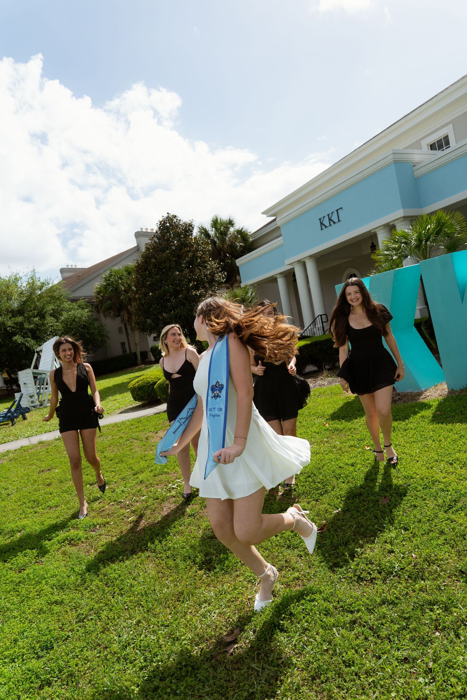 Young women in black dresses run across green lawn outside Kappa Kappa Gamma sorority house on sunny day.