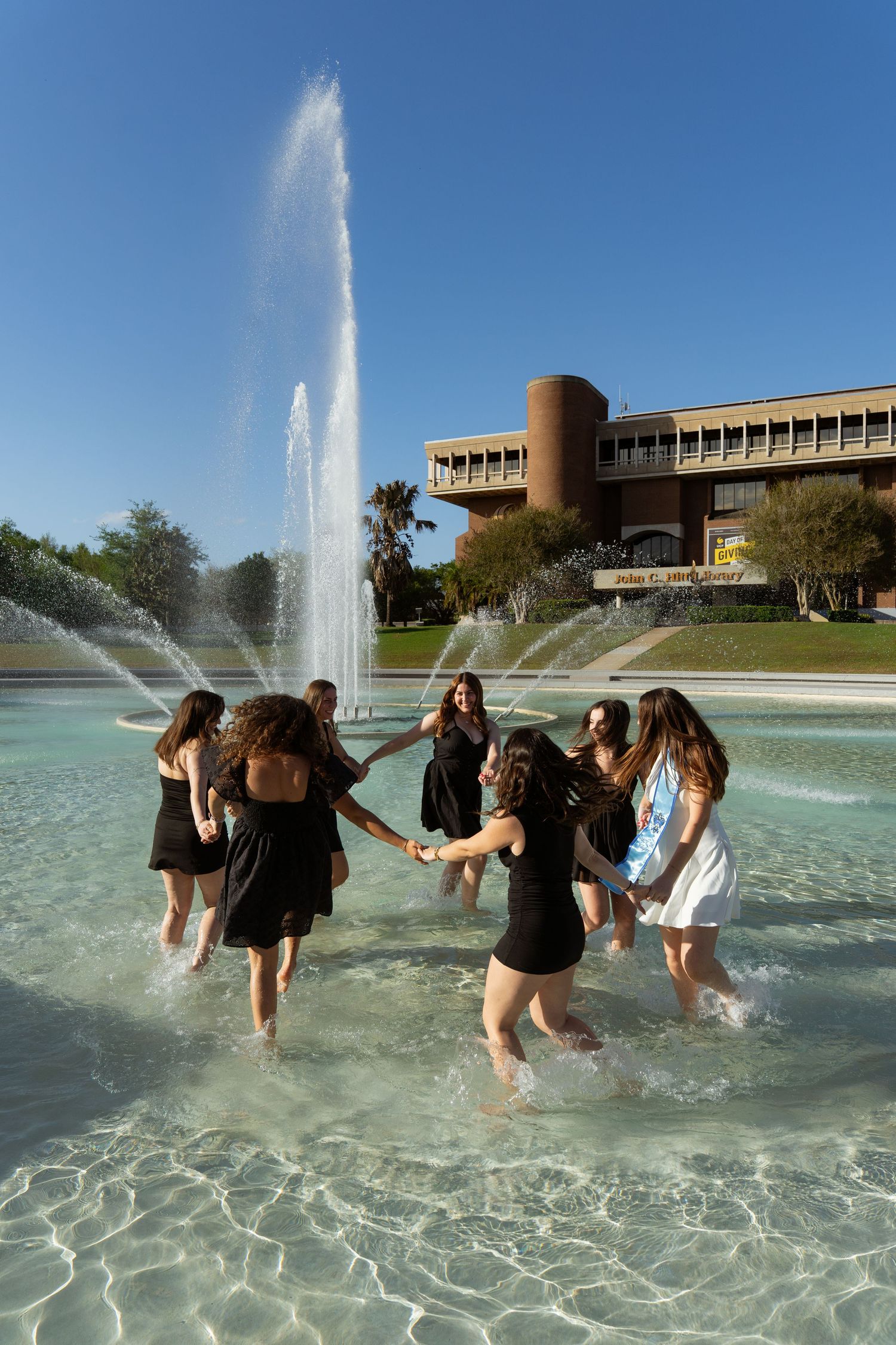 Group of young women dancing in a fountain outside a university building on a sunny day.