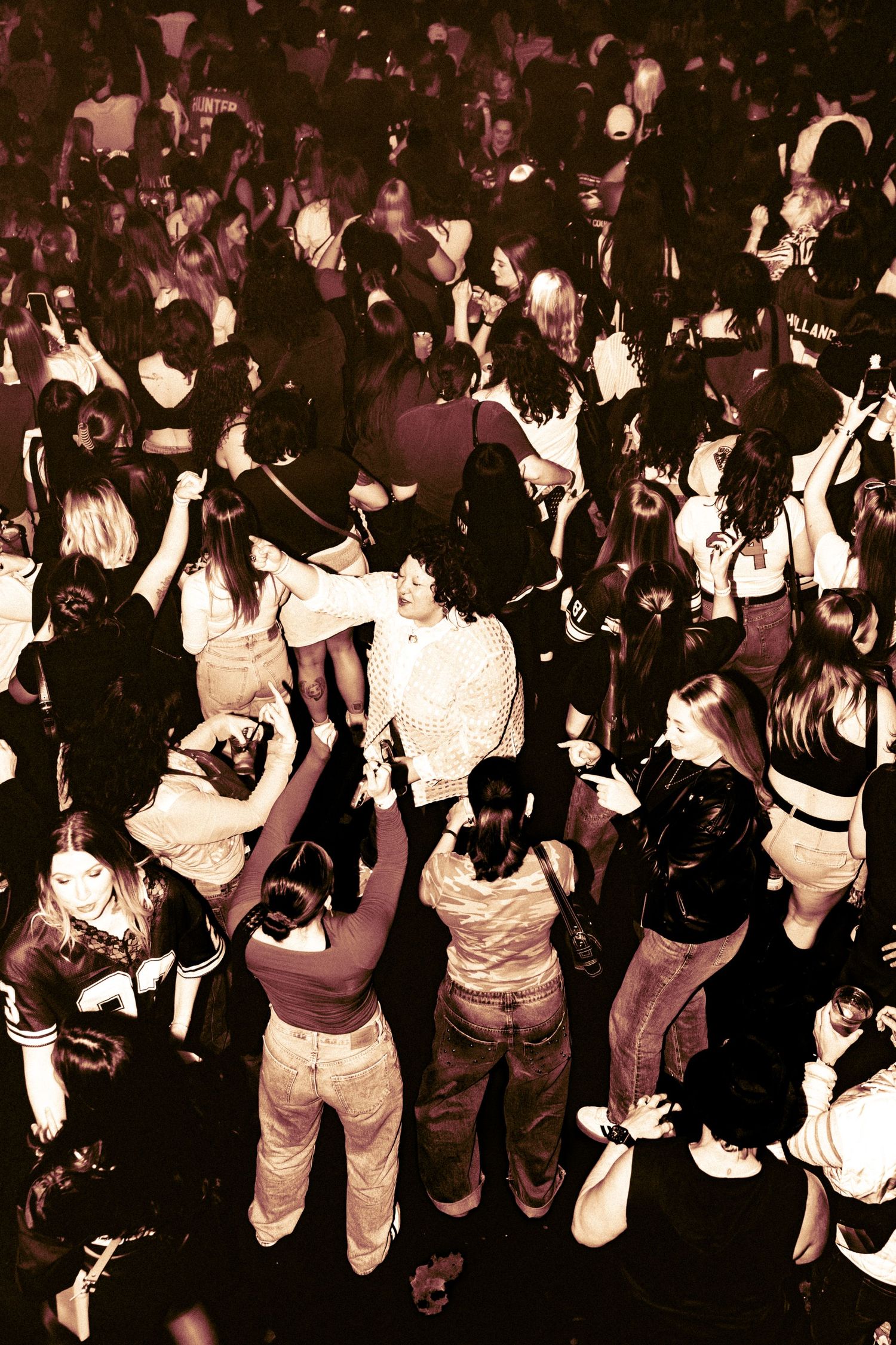 Aerial view of a large crowd of people dancing at a packed nightclub party under warm sepia-toned lighting.