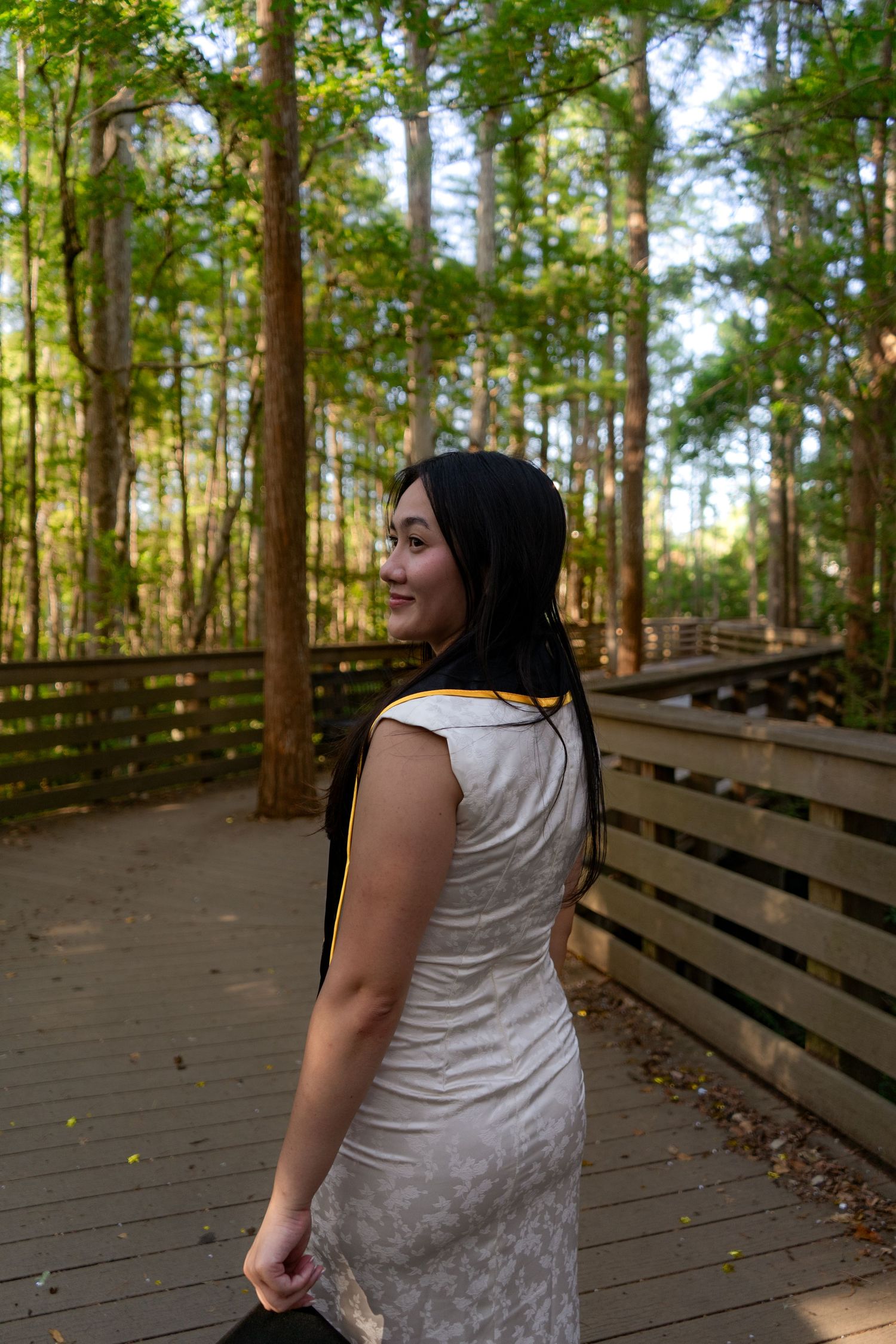 Young woman in white dress with graduation stole smiling on wooden boardwalk surrounded by tall forest trees.