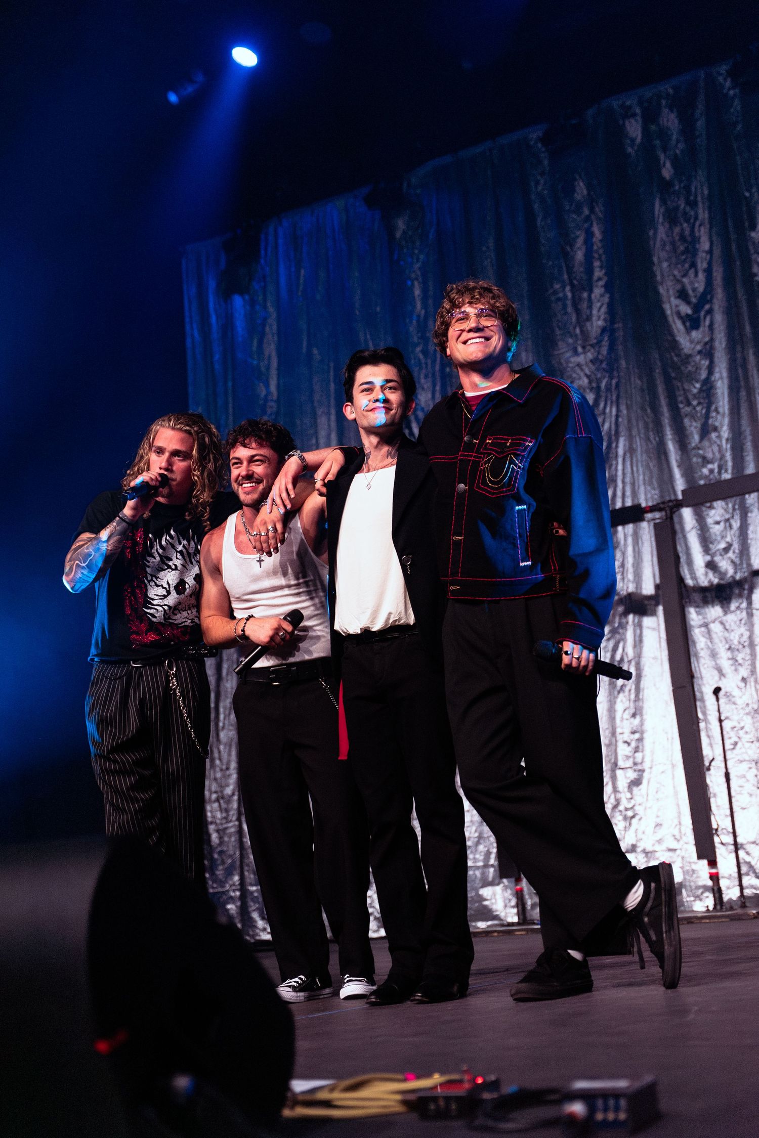 Four young male performers posing together on stage with blue lighting and silver curtain backdrop.