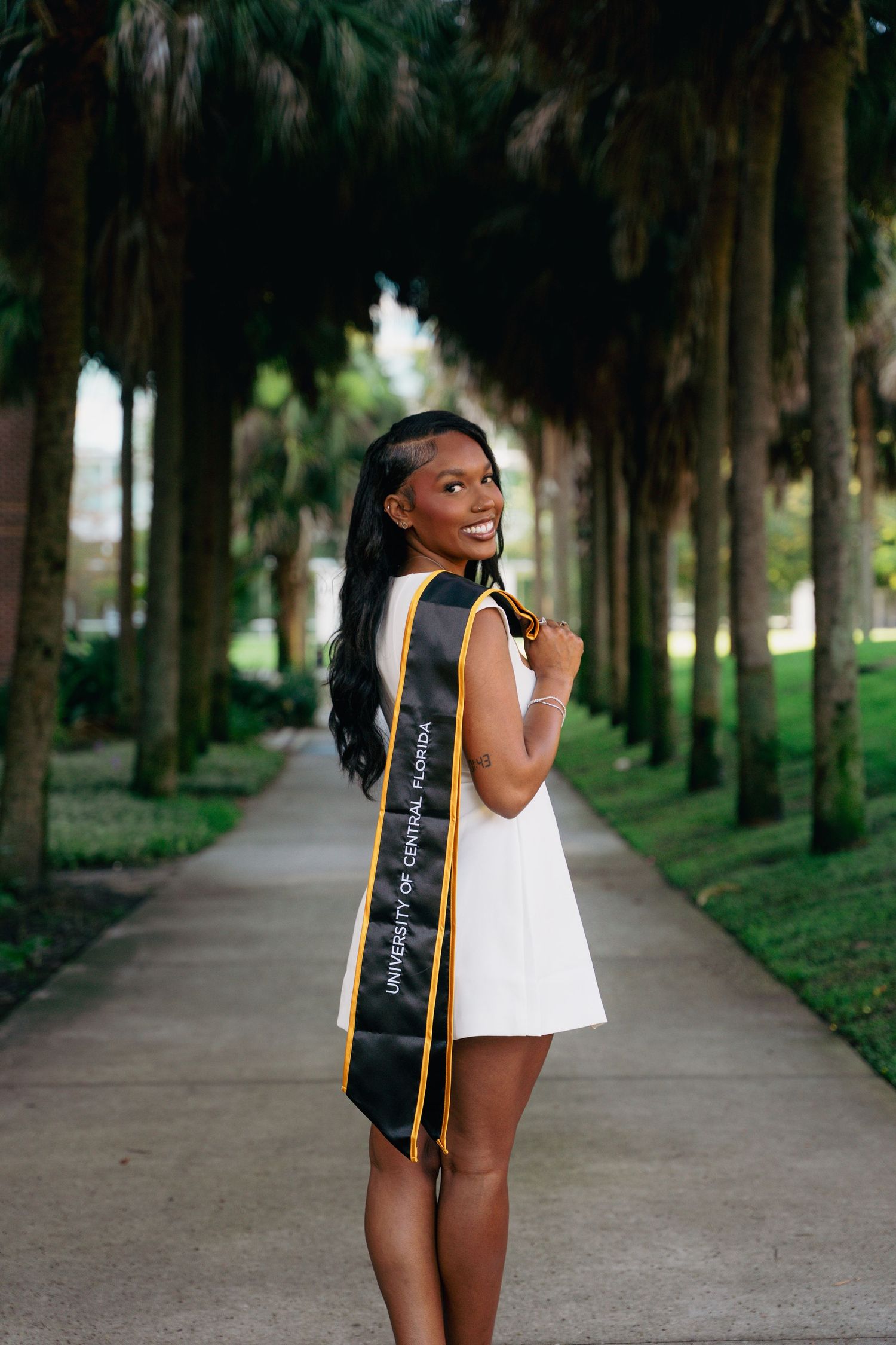 Smiling graduate in white dress wearing University of Central Florida black and gold sash outdoors.