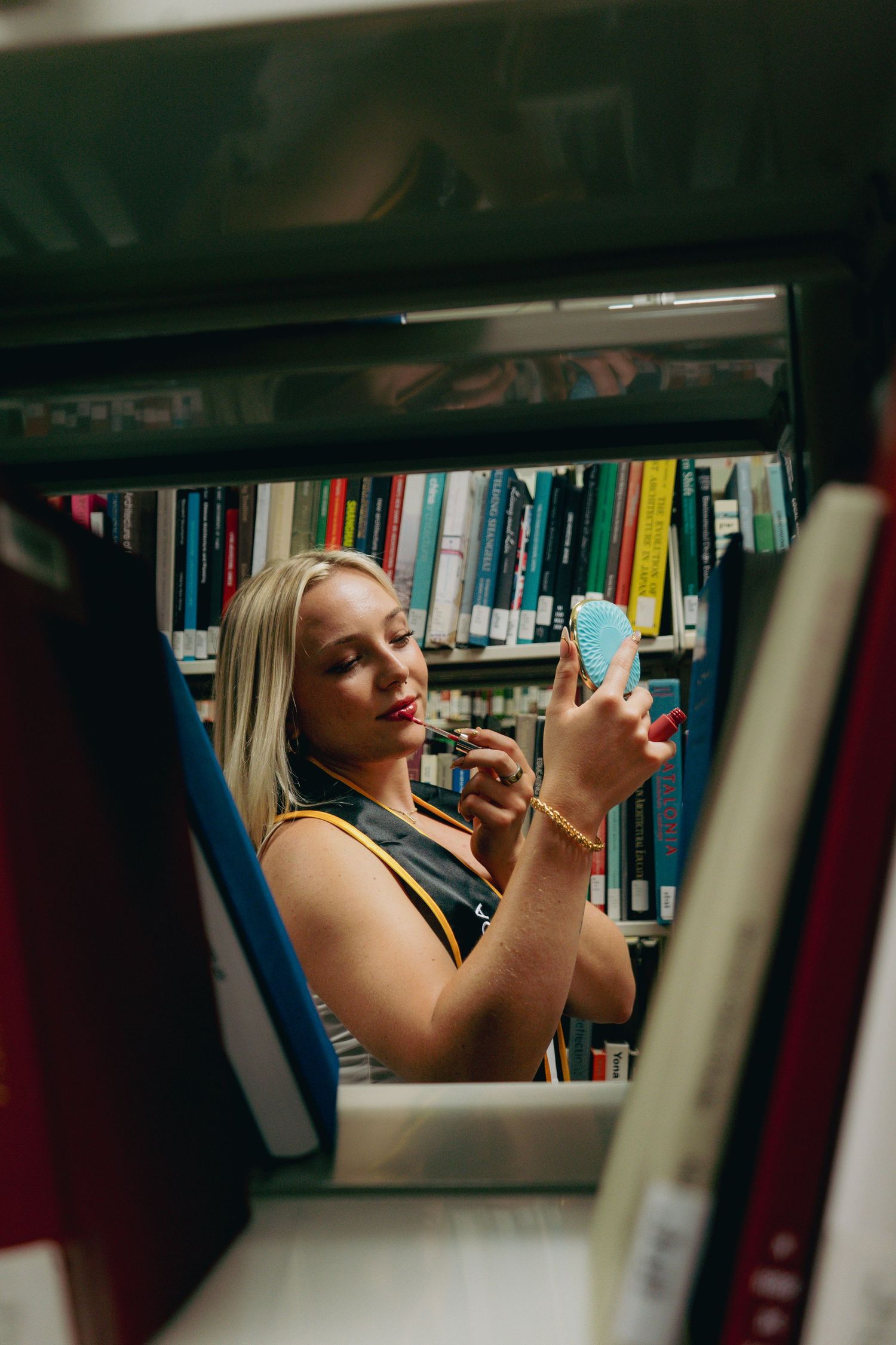 Blonde woman applies lipstick using compact mirror between colorful library bookshelves wearing graduation stole.