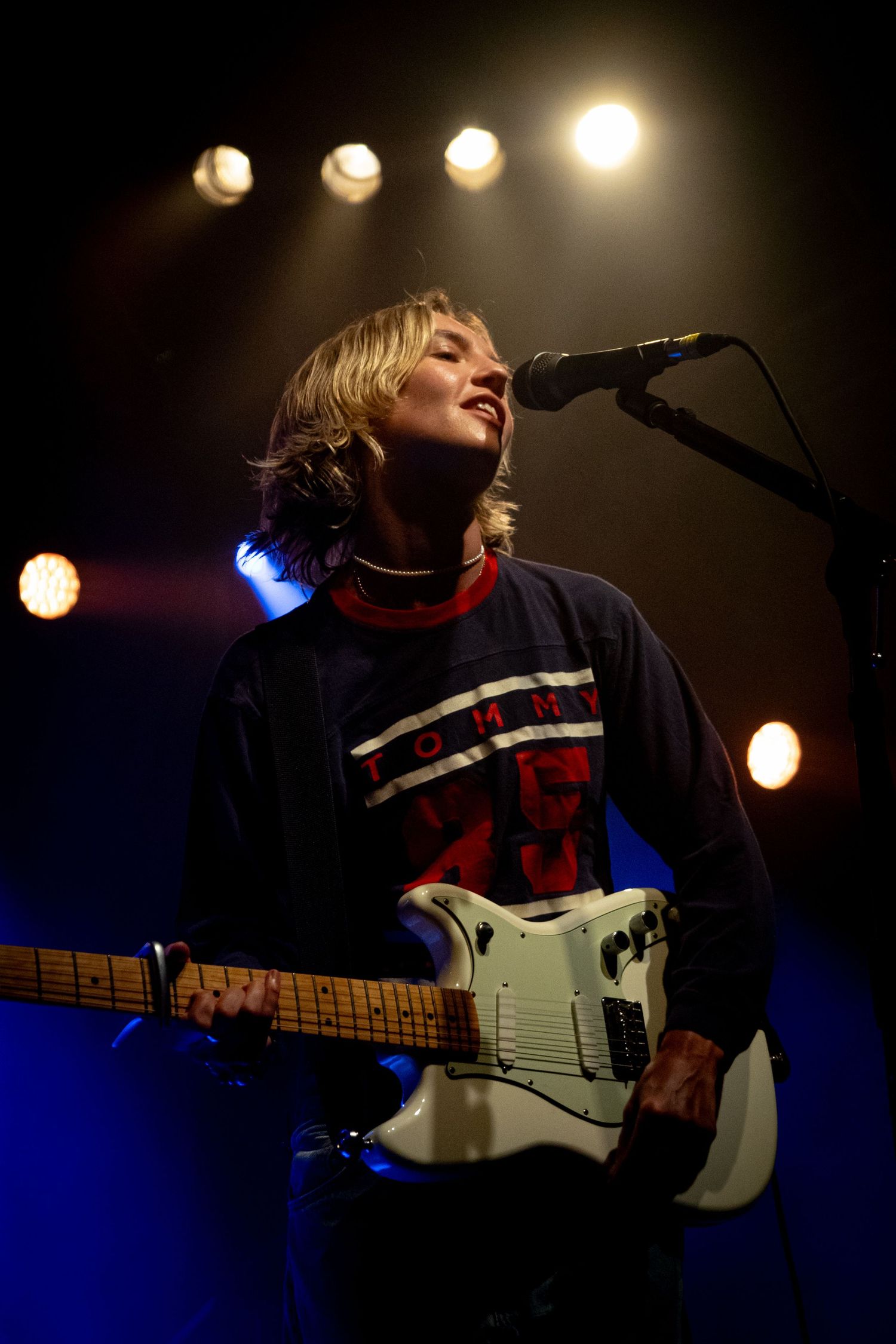 Blonde musician performing on stage with a white electric guitar under warm stage lights.