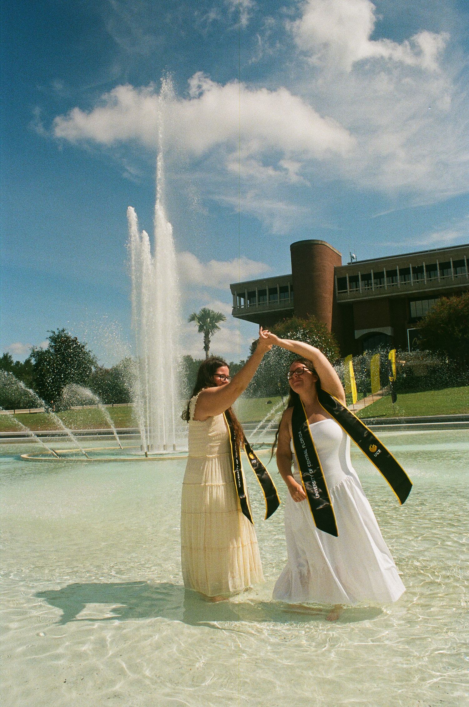 Two female graduates in white dresses and black sashes celebrate in a fountain with a building and blue sky behind them.