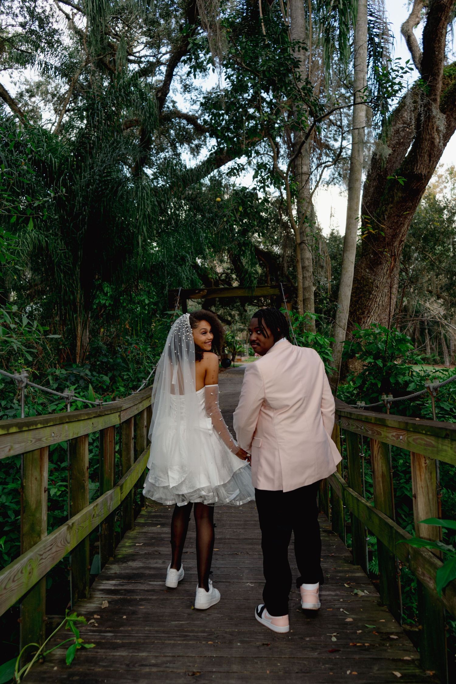 Bride in short lace wedding dress and veil walks hand-in-hand with groom in pink blazer on bridge.