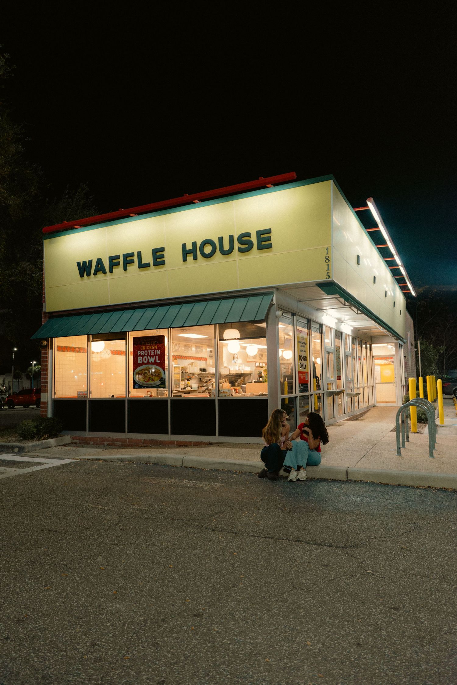 Illuminated Waffle House restaurant exterior at night with two people sitting outside on the sidewalk.