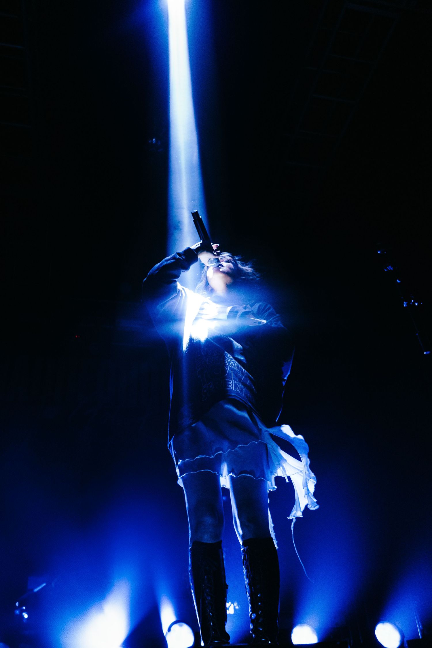 Solo female performer stands bathed in dramatic blue beam of light on dark concert stage.