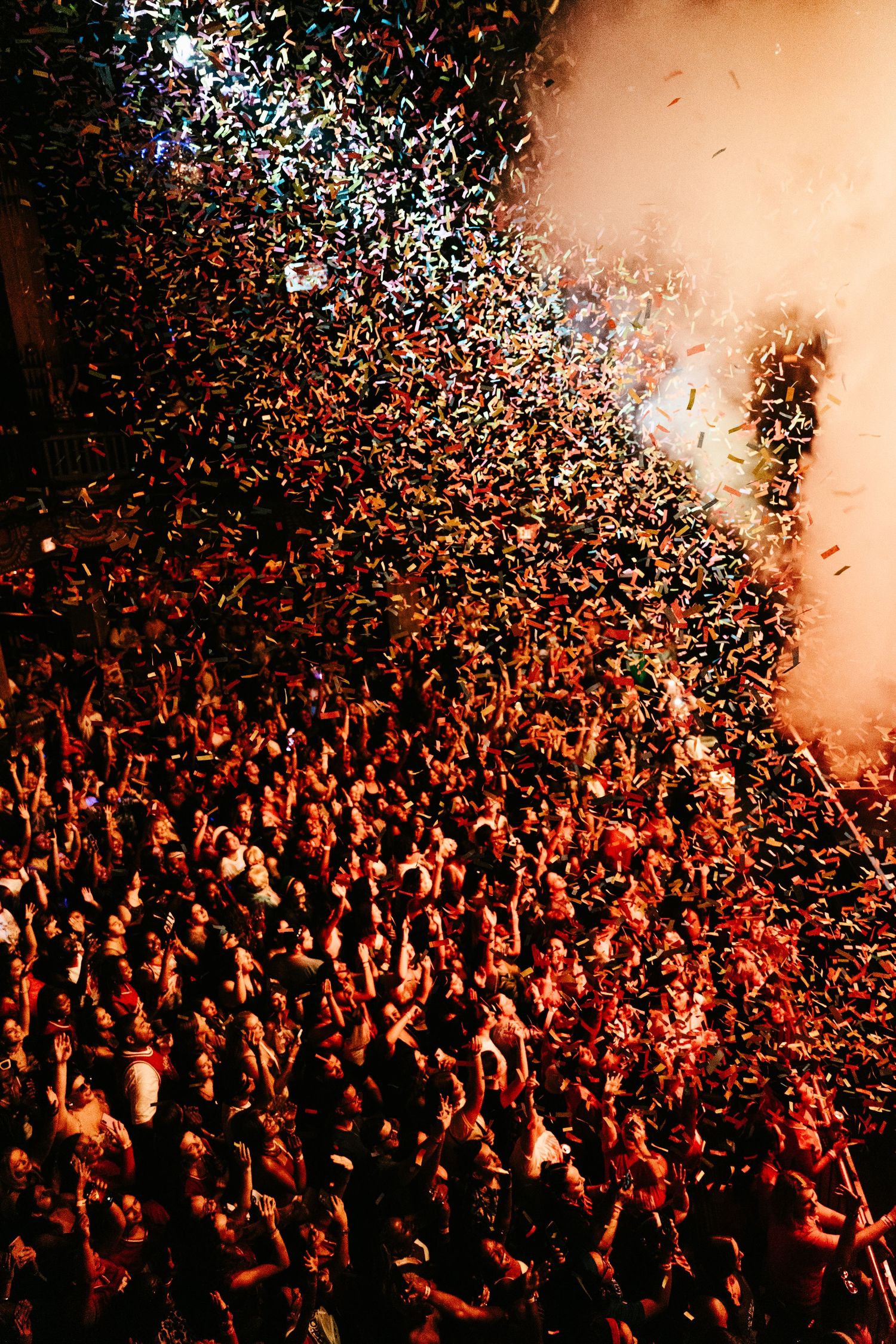 Aerial view of concert crowd showered in multicolored confetti under bright stage fog and lights.