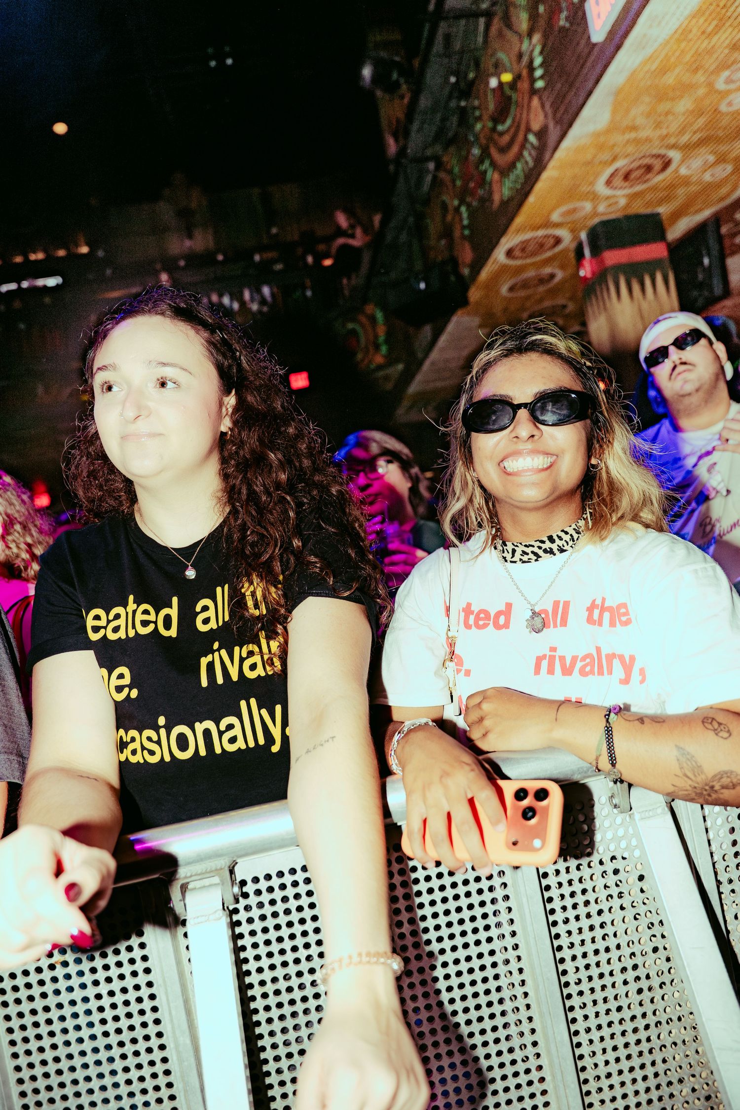 Two smiling women in matching black and white graphic tees lean on a concert barrier at a live music event.
