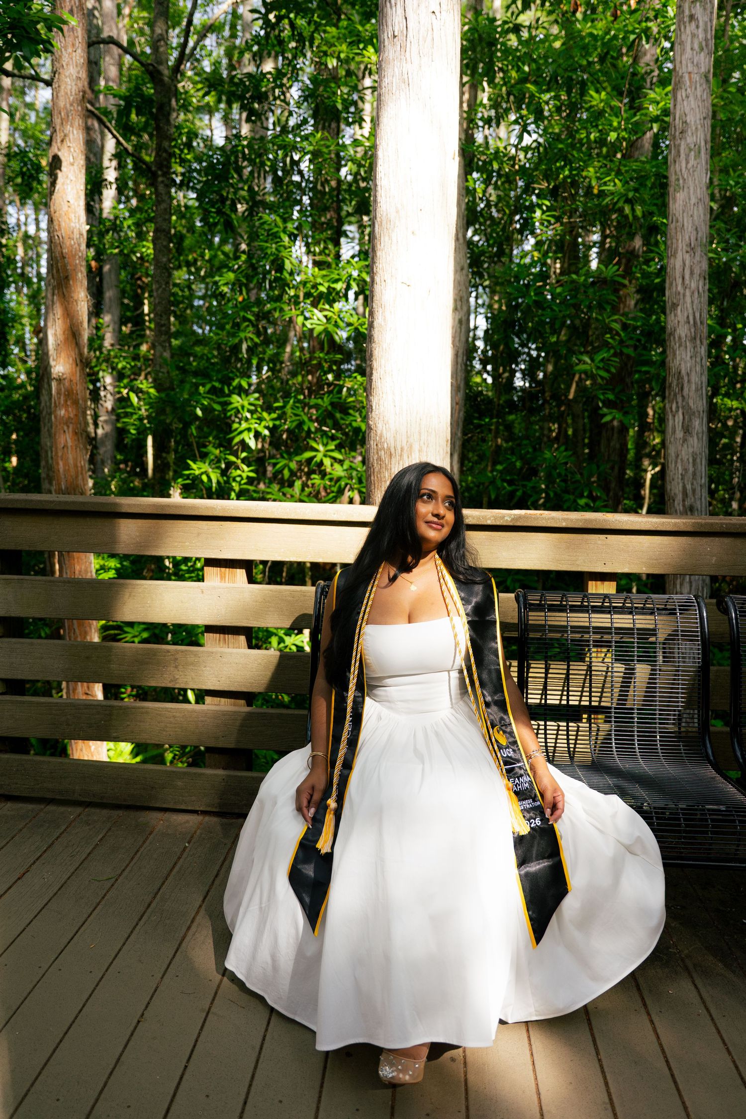 Young woman in white dress and graduation regalia poses on wooden deck surrounded by lush green trees.
