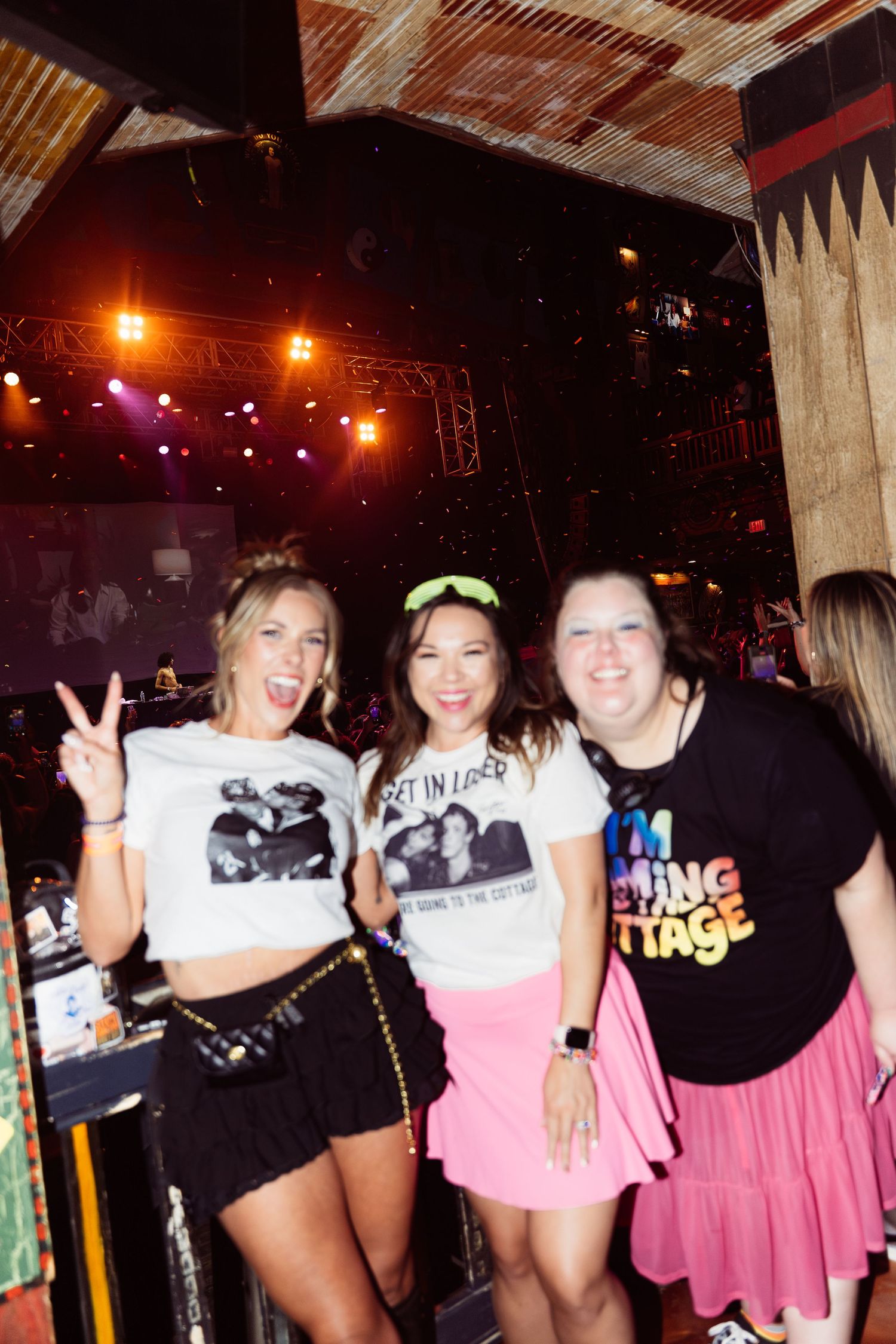 Three women smiling and posing at a concert venue, wearing fun graphic tees with stage lights in background.