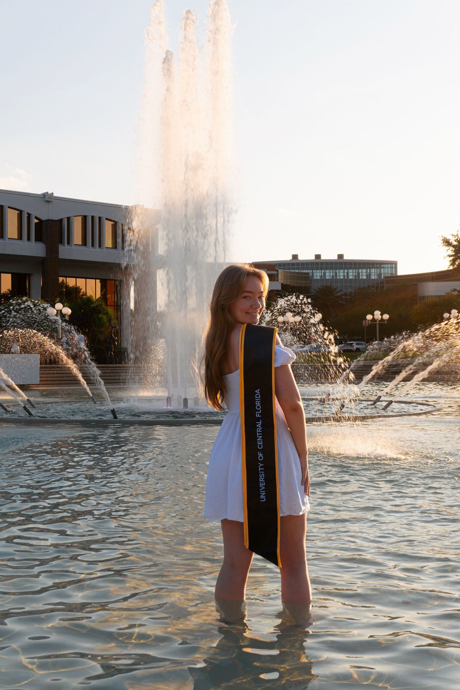 Young woman in white dress holding graduation sash standing in fountain at golden hour.