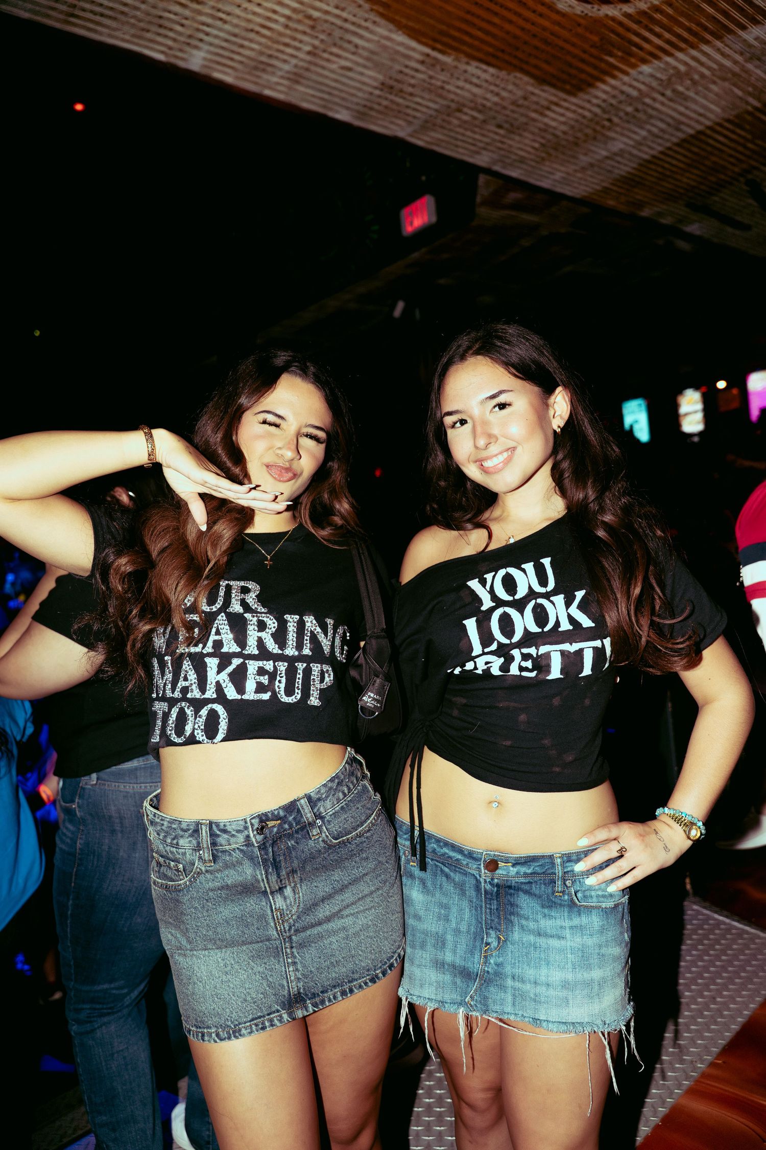 Two young women pose in black graphic crop tees and denim skirts at a nightclub event.