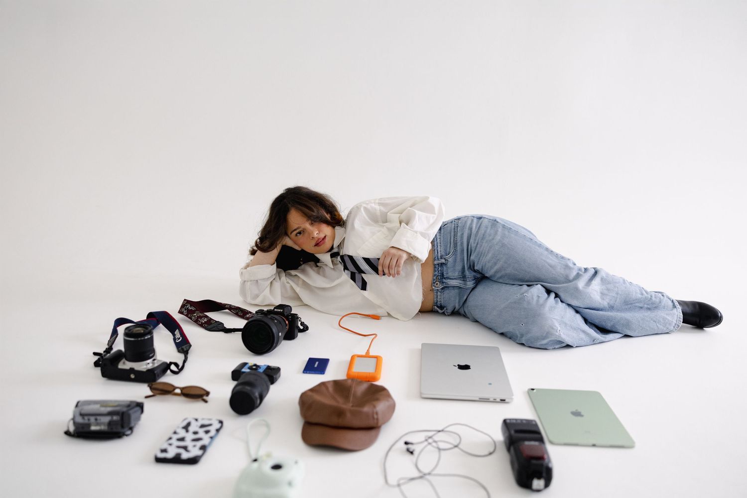 Photographer lying on white floor surrounded by camera gear, tablet, phone, and accessories.
