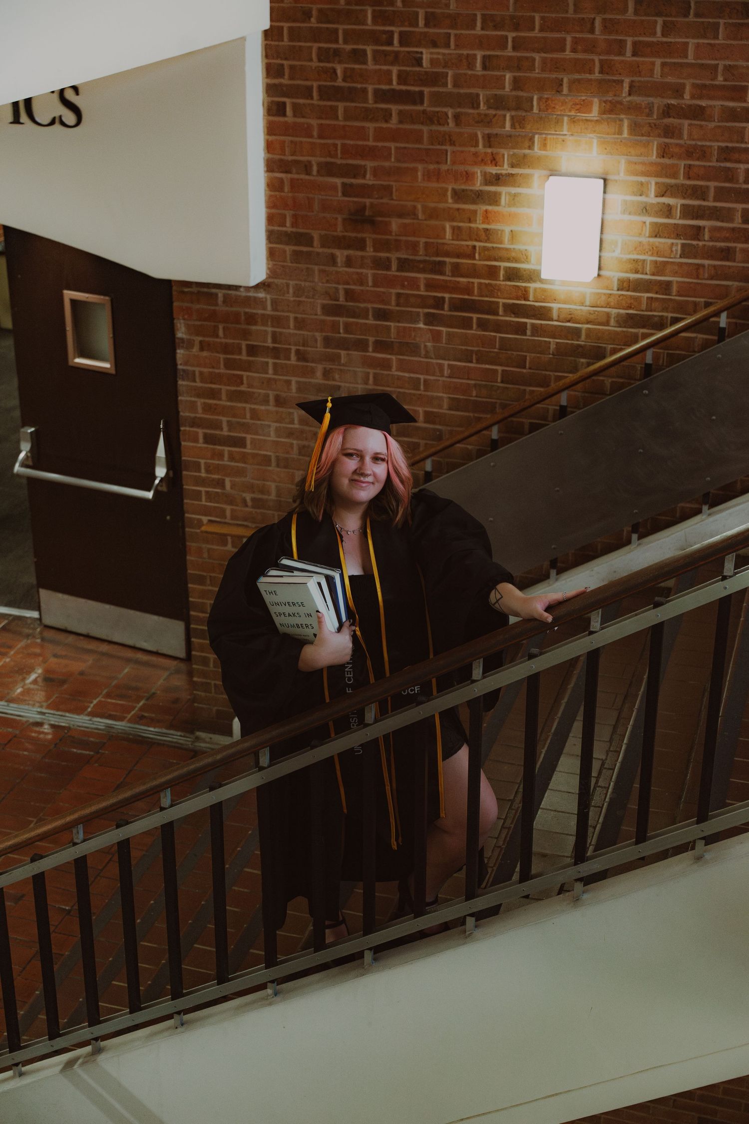 Smiling graduate in black cap and gown holds books on a staircase inside a brick university building.