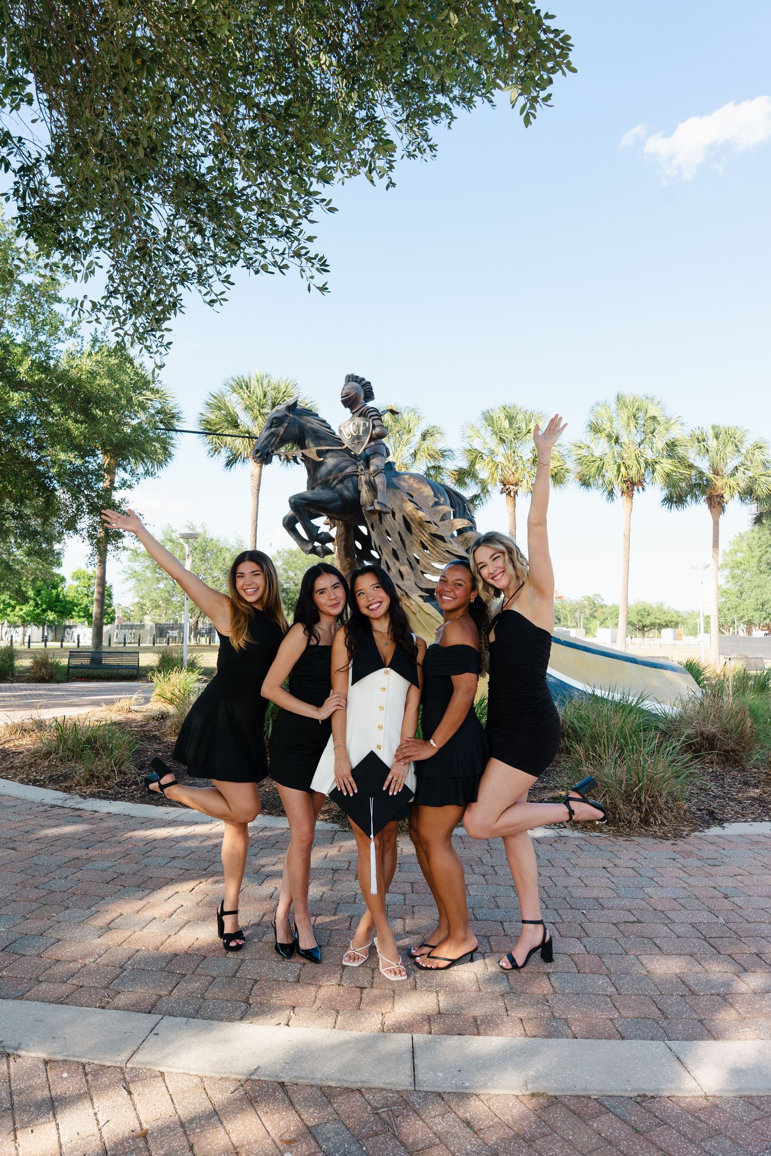 Five young women in black dresses pose joyfully in front of a bronze equestrian statue outdoors.