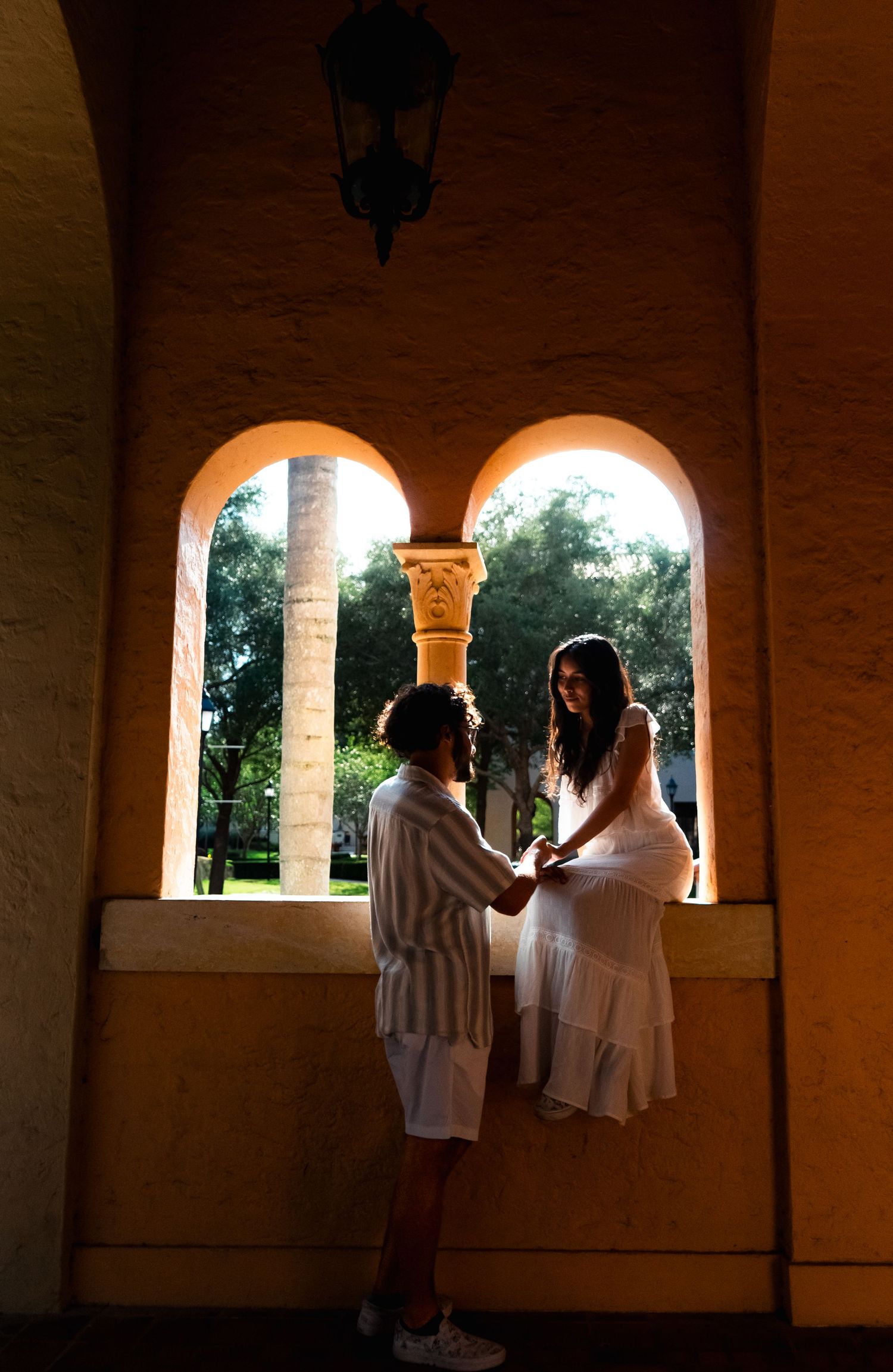 Romantic couple holding hands in a sunlit arched window alcove of a historic building.