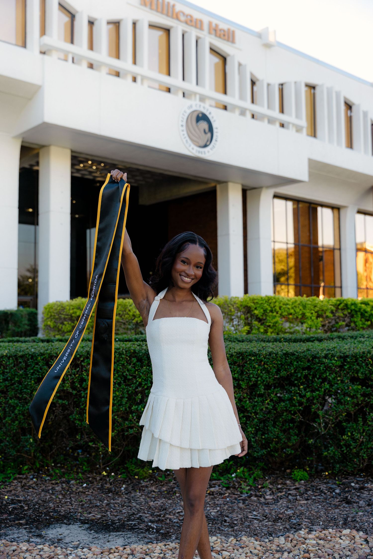 Young woman in white dress holds UCF black and gold graduation sash outside Millican Hall.