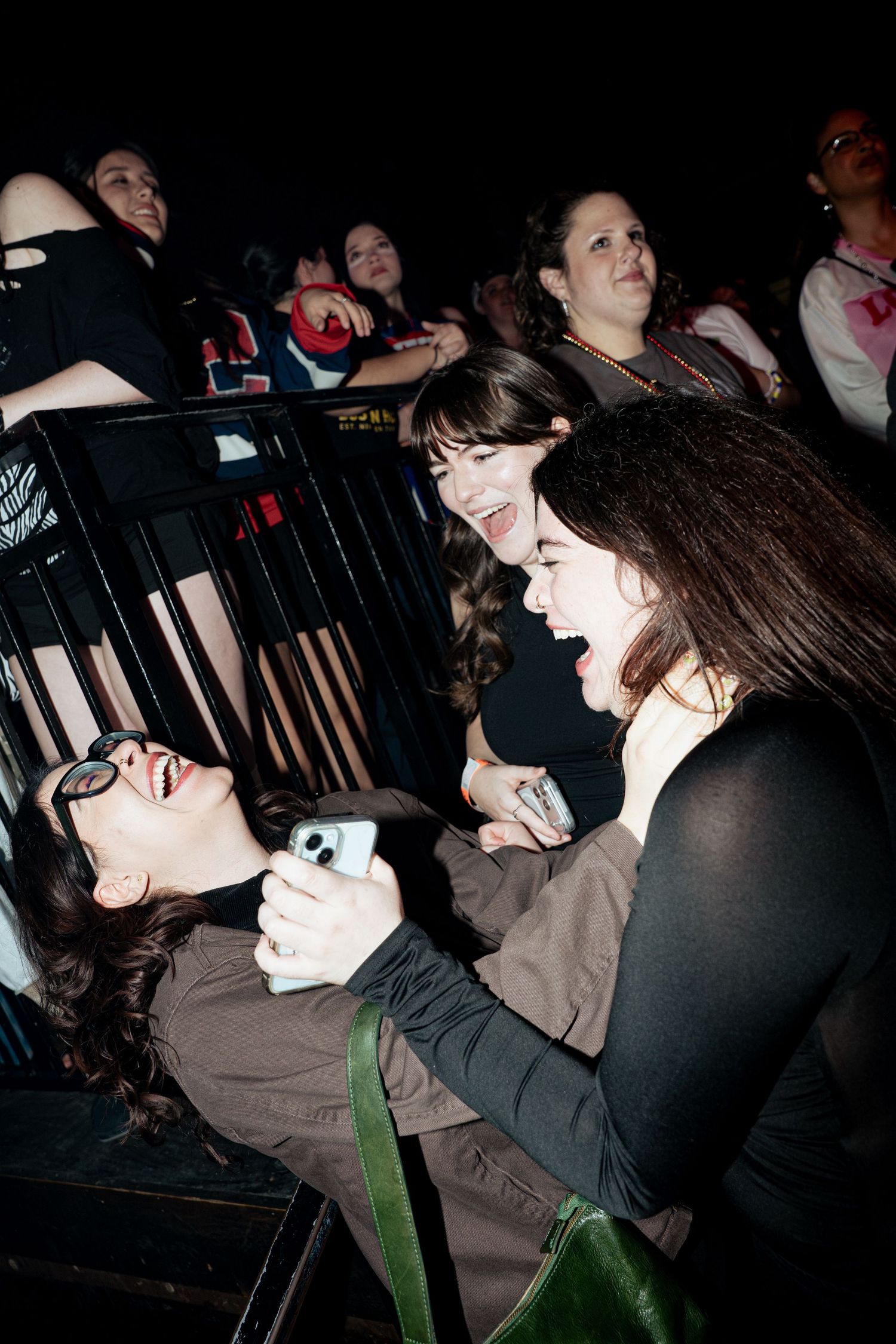 Three young women laughing and taking selfies together at a dark concert venue barrier.