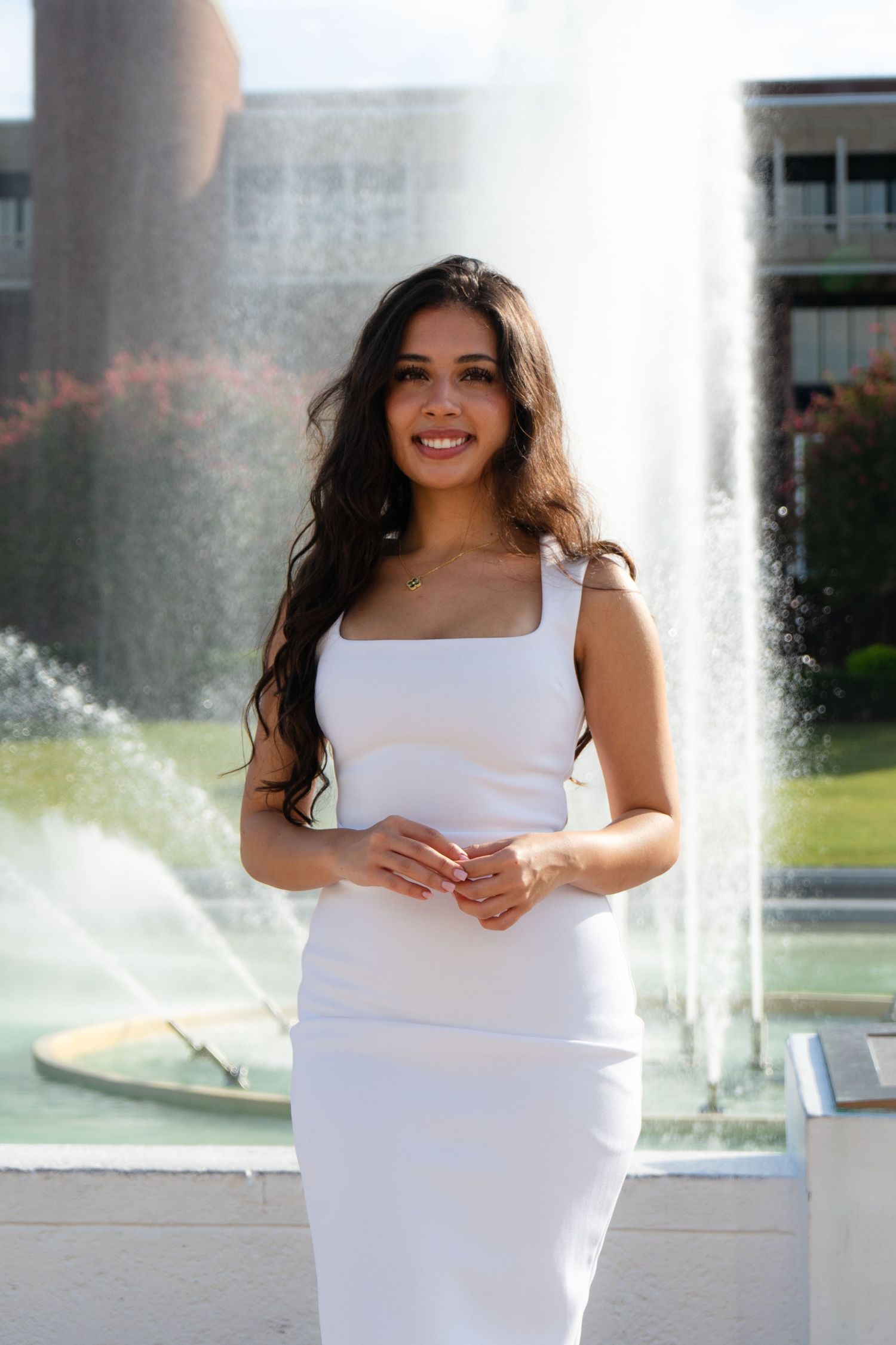 Smiling young woman in white midi dress standing before campus fountain on sunny day.