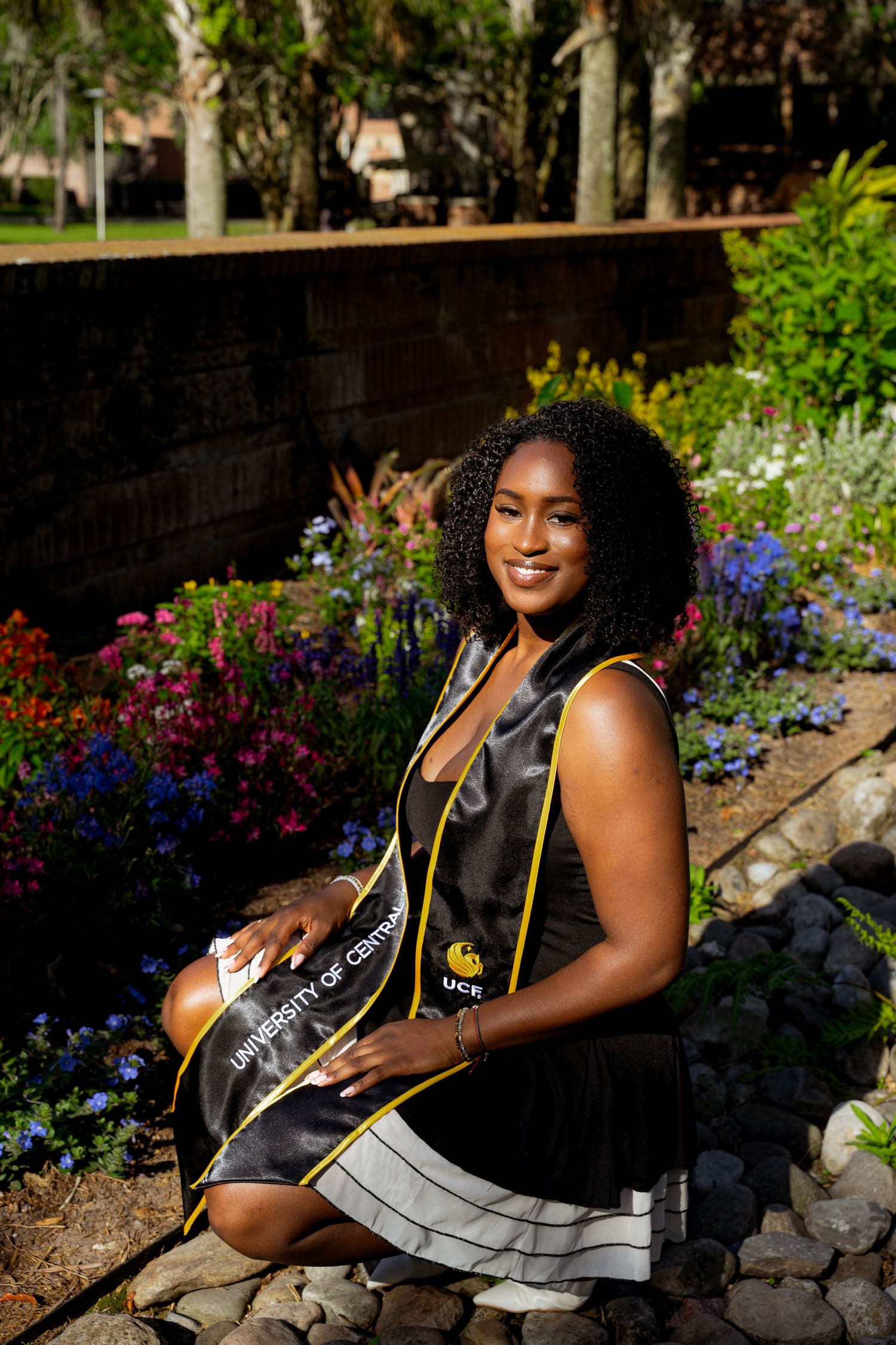 A smiling woman in UCF graduation regalia poses among colorful flowers on a sunny day.