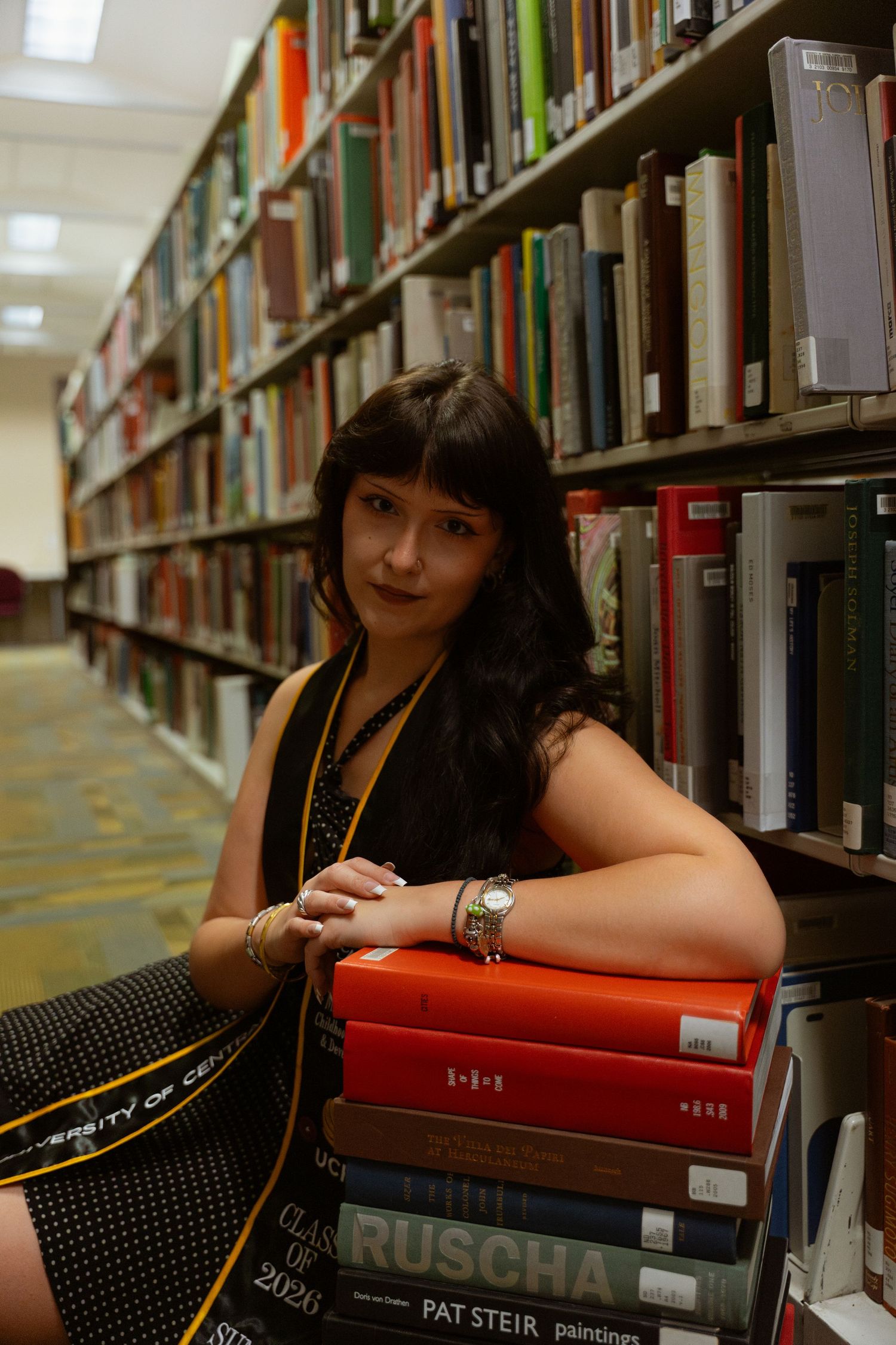 Woman with dark hair leans on stack of books in library aisle, shelves of books visible behind her.