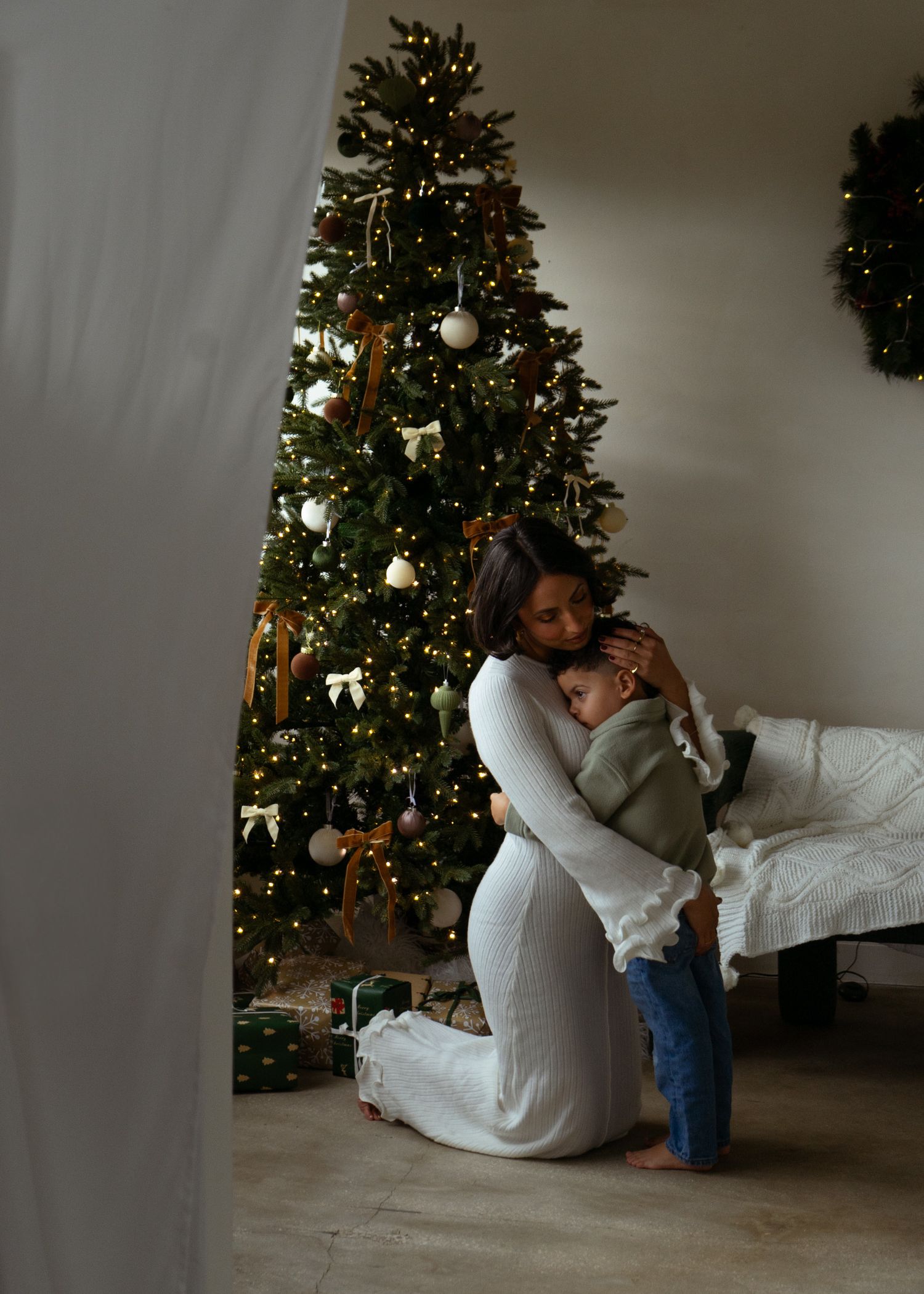 Mother kneeling and embracing her young child in front of a beautifully lit Christmas tree.