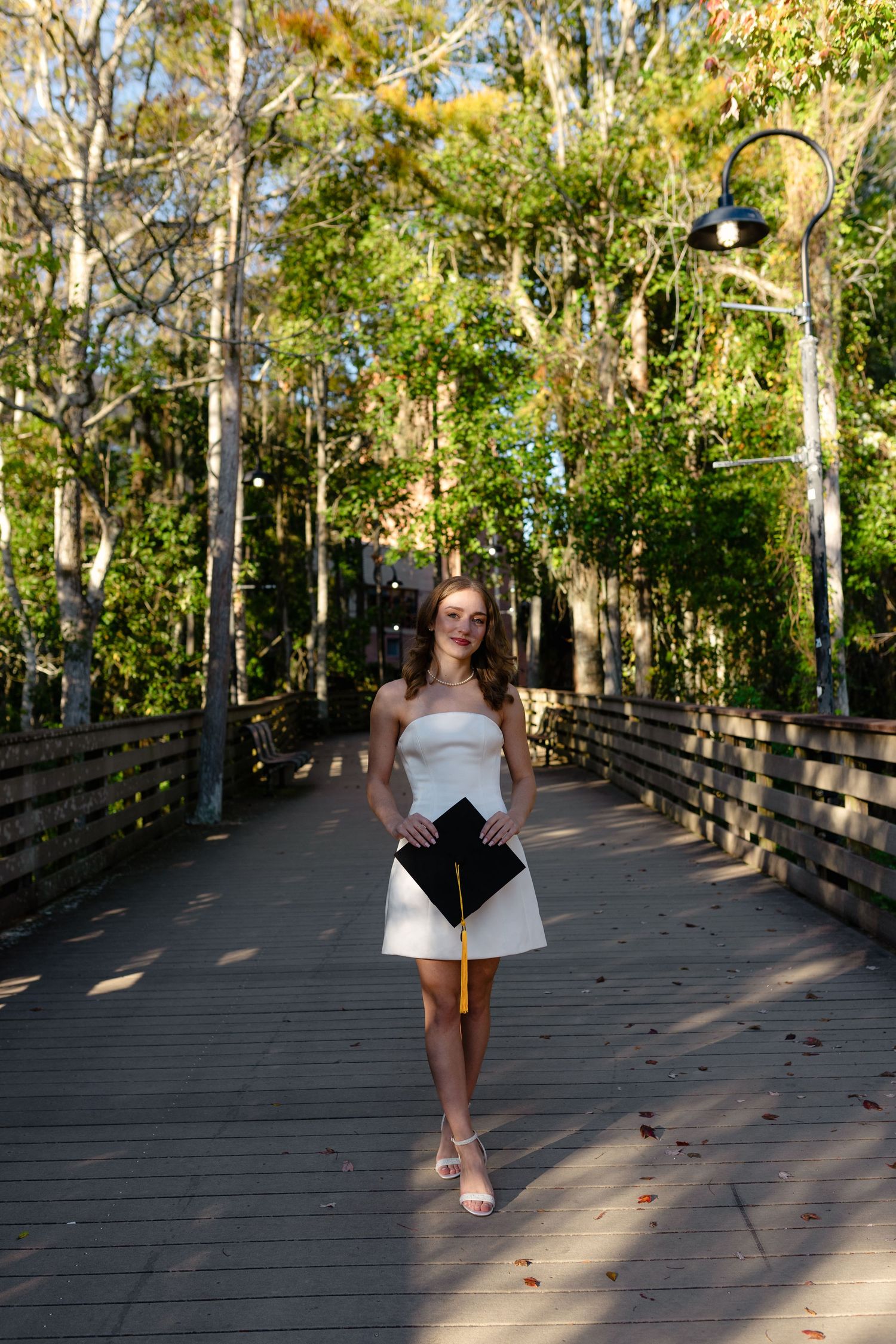 Young woman in white dress holding graduation cap on a sunlit forest boardwalk.