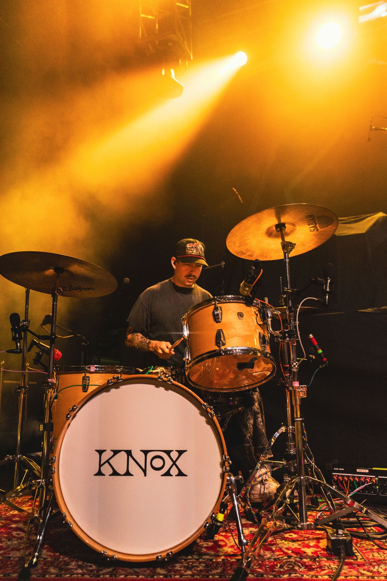 Drummer performing on Knox drum kit under dramatic golden stage lighting with smoke effects.