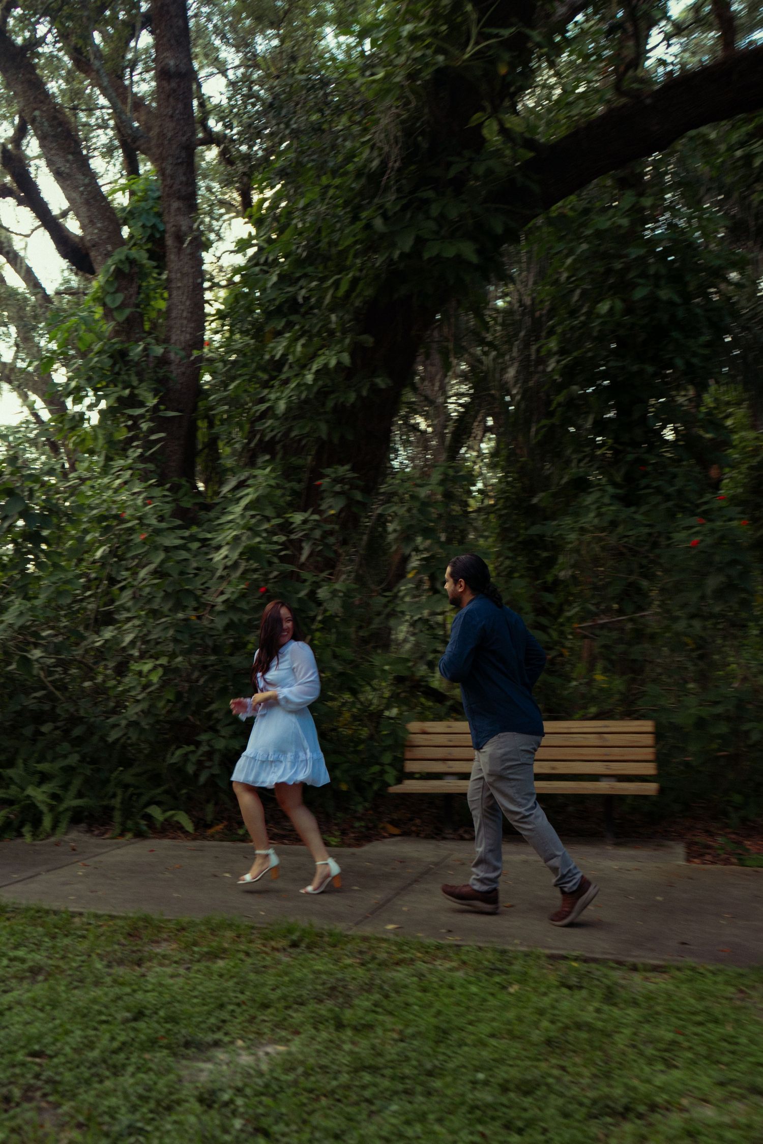 Couple walking together on a shaded garden path surrounded by lush green trees and a wooden bench.