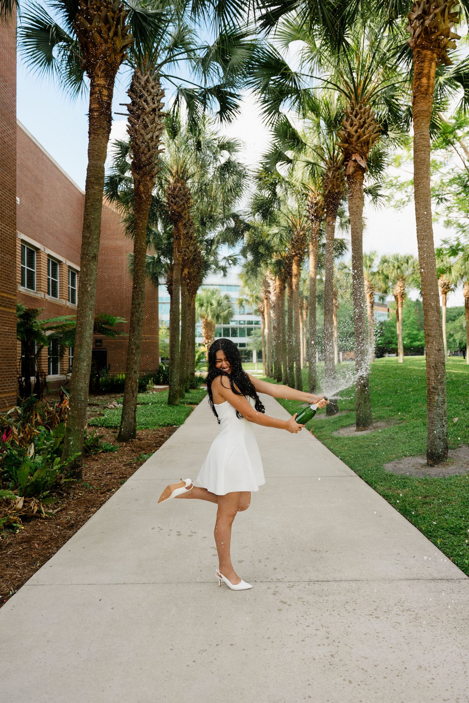 Young woman in white dress celebrating outdoors, spraying champagne among palm trees on a sunny campus walkway.
