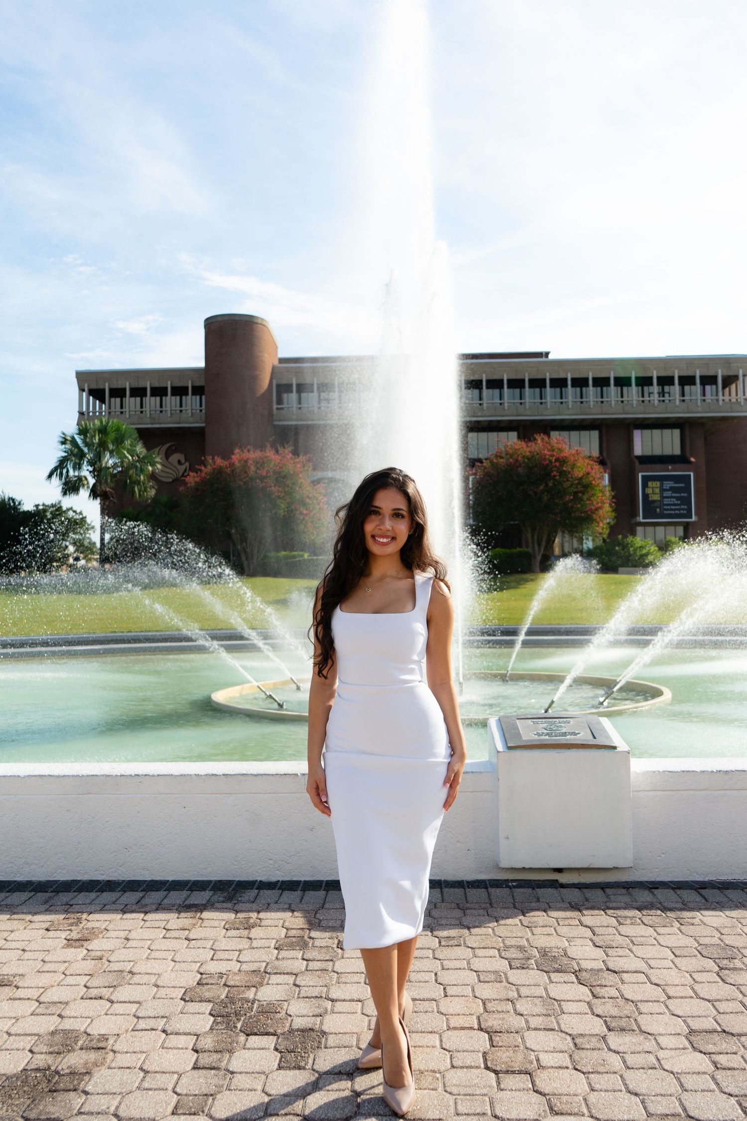 Woman in white midi dress posing in front of a large fountain with a brick building backdrop on a sunny day.