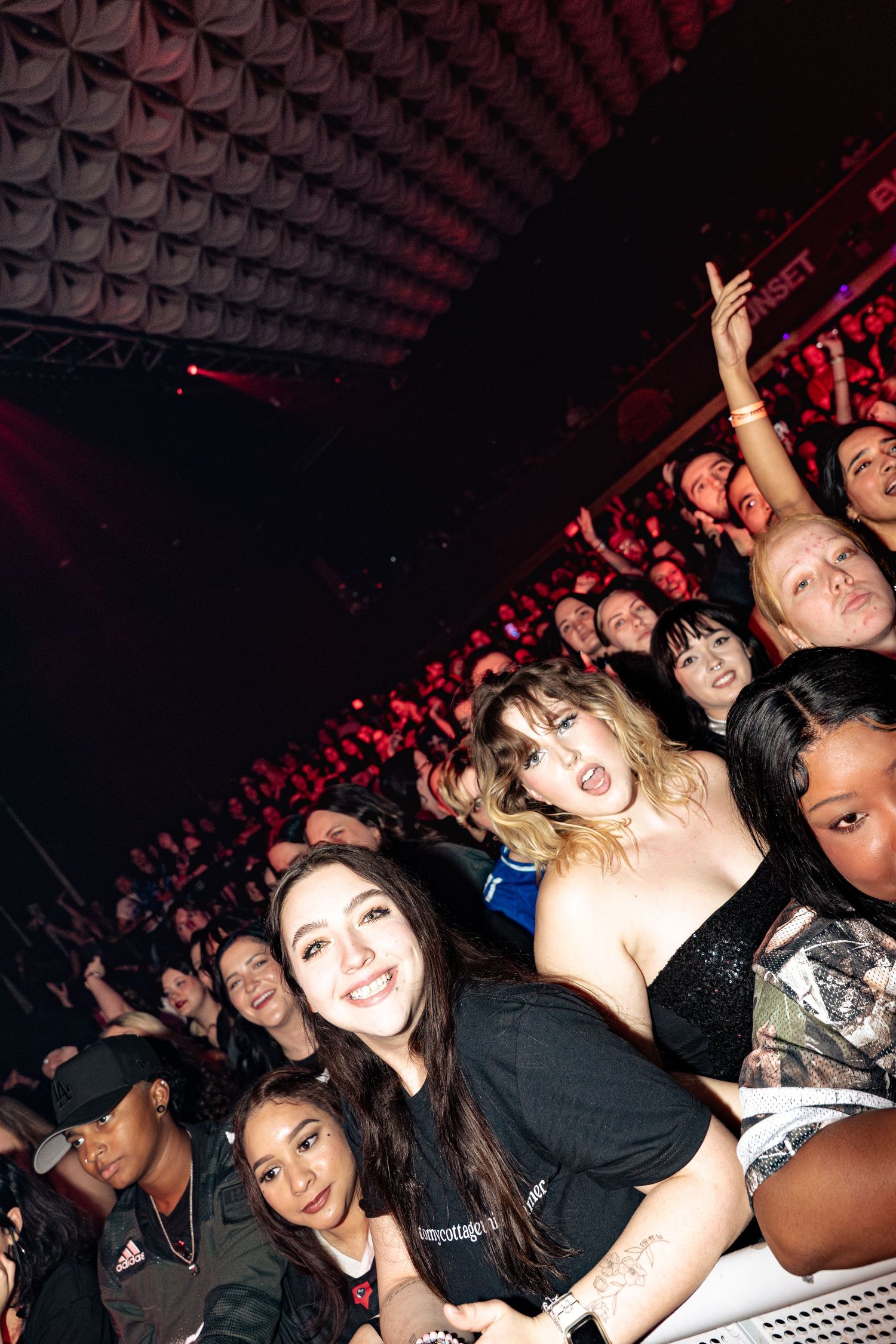 Excited crowd of fans at a live music concert venue with red stage lighting.