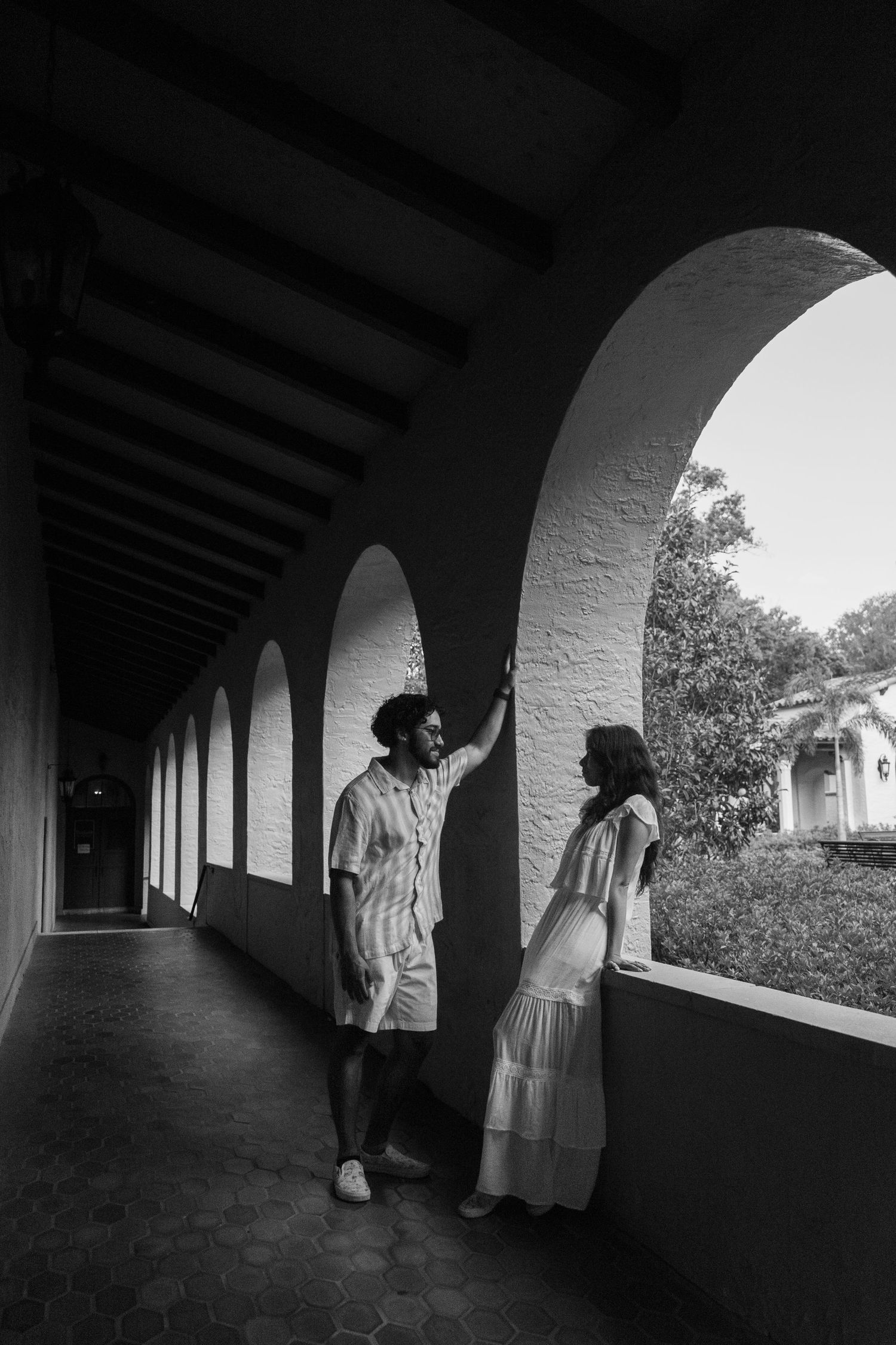 Black and white photo of couple standing under arched colonnade in casual white outfits.