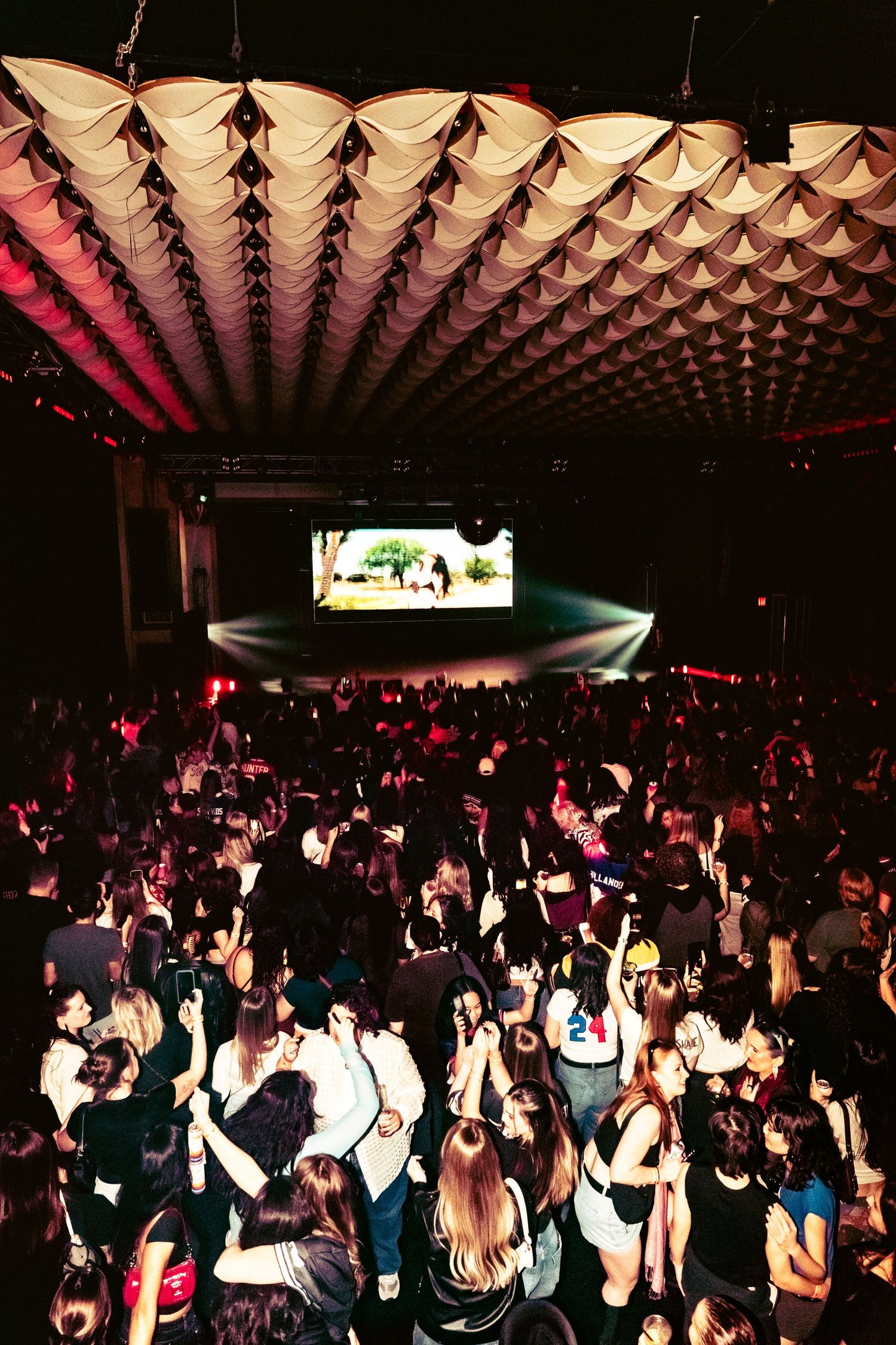 Packed nightclub crowd dancing under decorative ceiling with stage screen glowing in background.