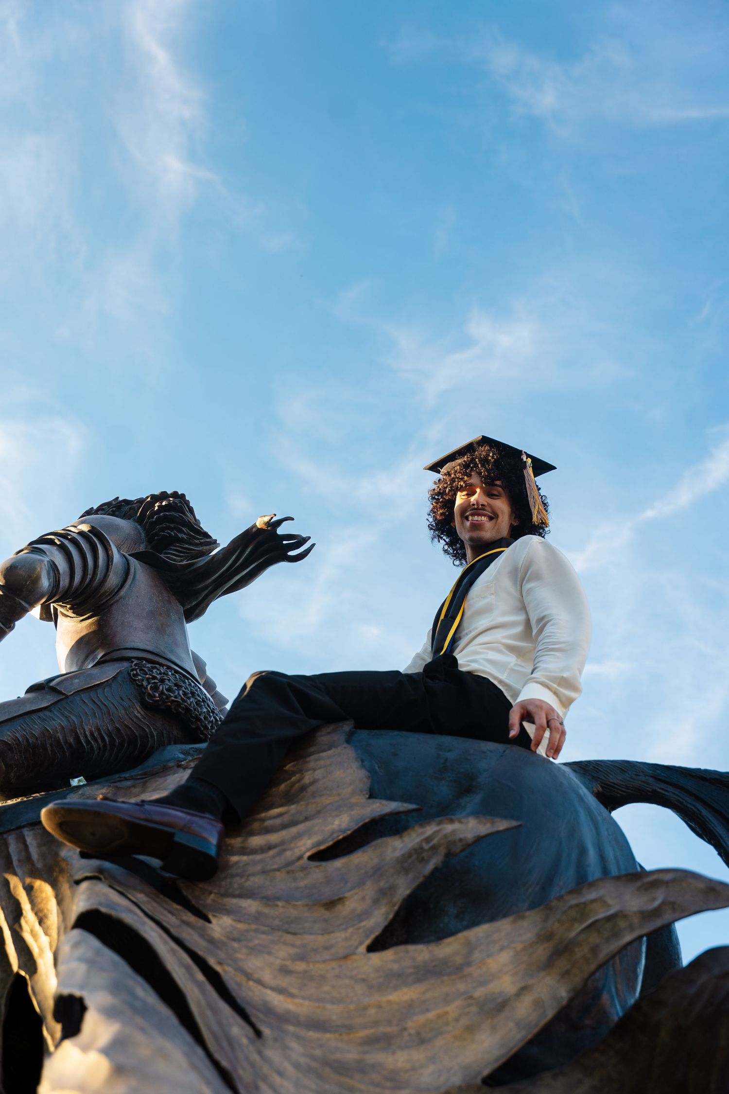 Graduate in cap and gown sitting atop a bronze equestrian statue against a blue sky.