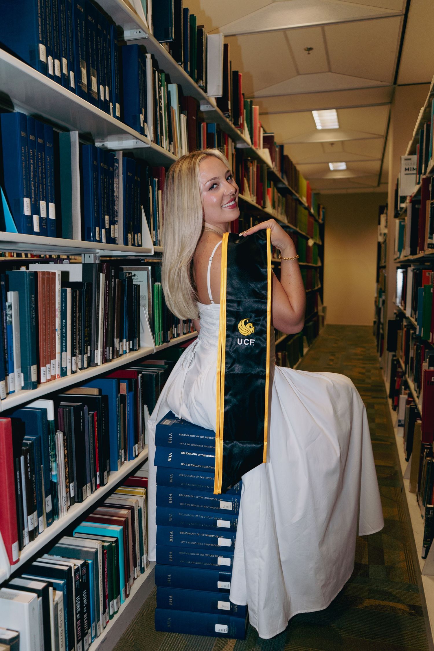 Blonde UCF graduate in white dress displays gold and black graduation stole between library bookshelves.