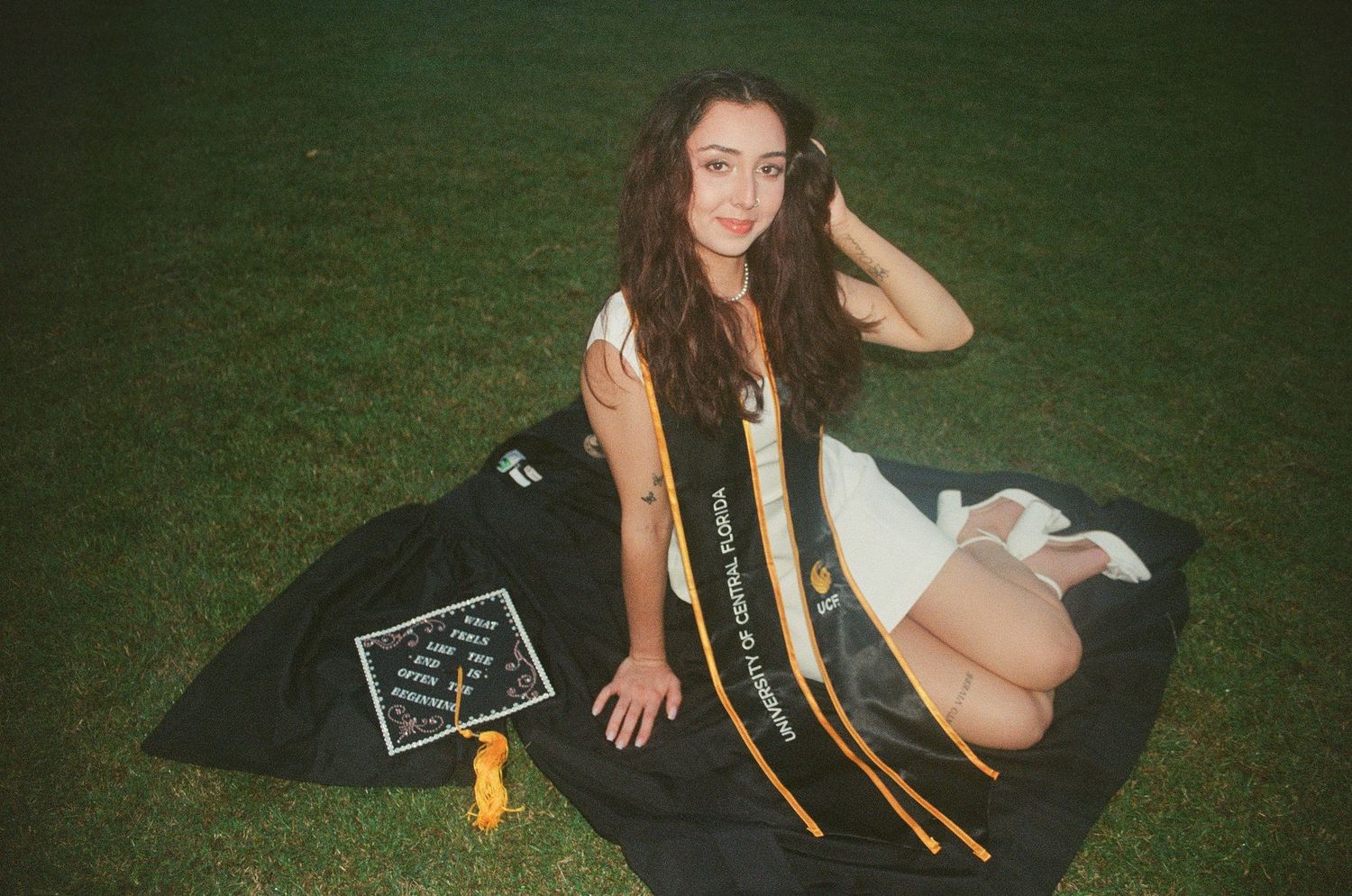 Young woman in graduation gown and UCF stole poses on green grass with decorated cap.