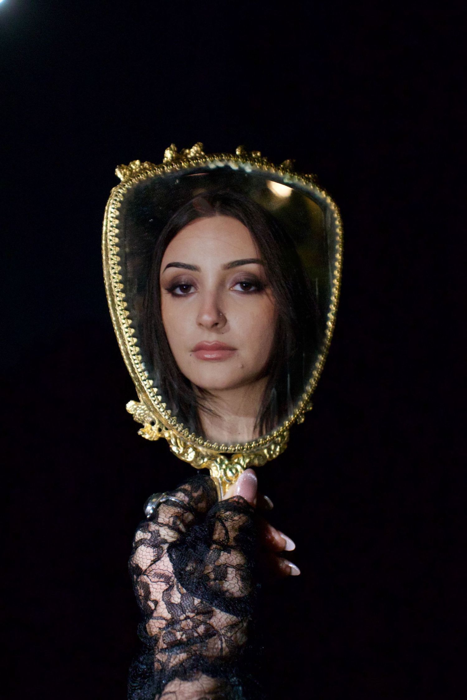 Young woman with dark makeup reflected in ornate gold hand mirror against black background.