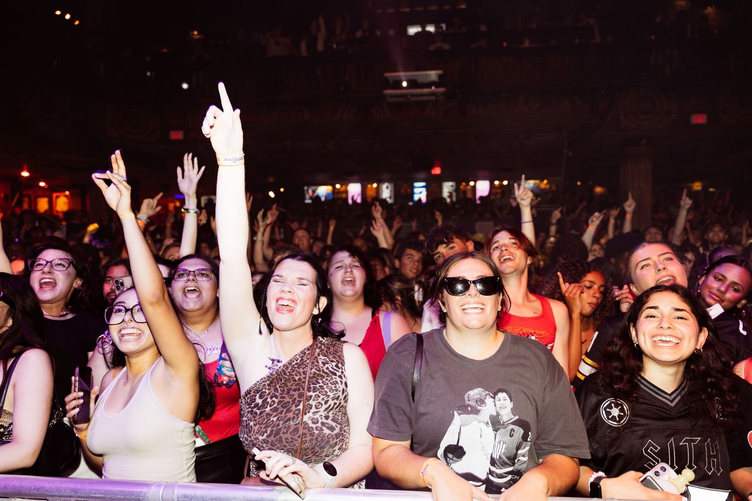 Excited concert crowd cheering and raising hands at a live music event in a packed indoor venue.