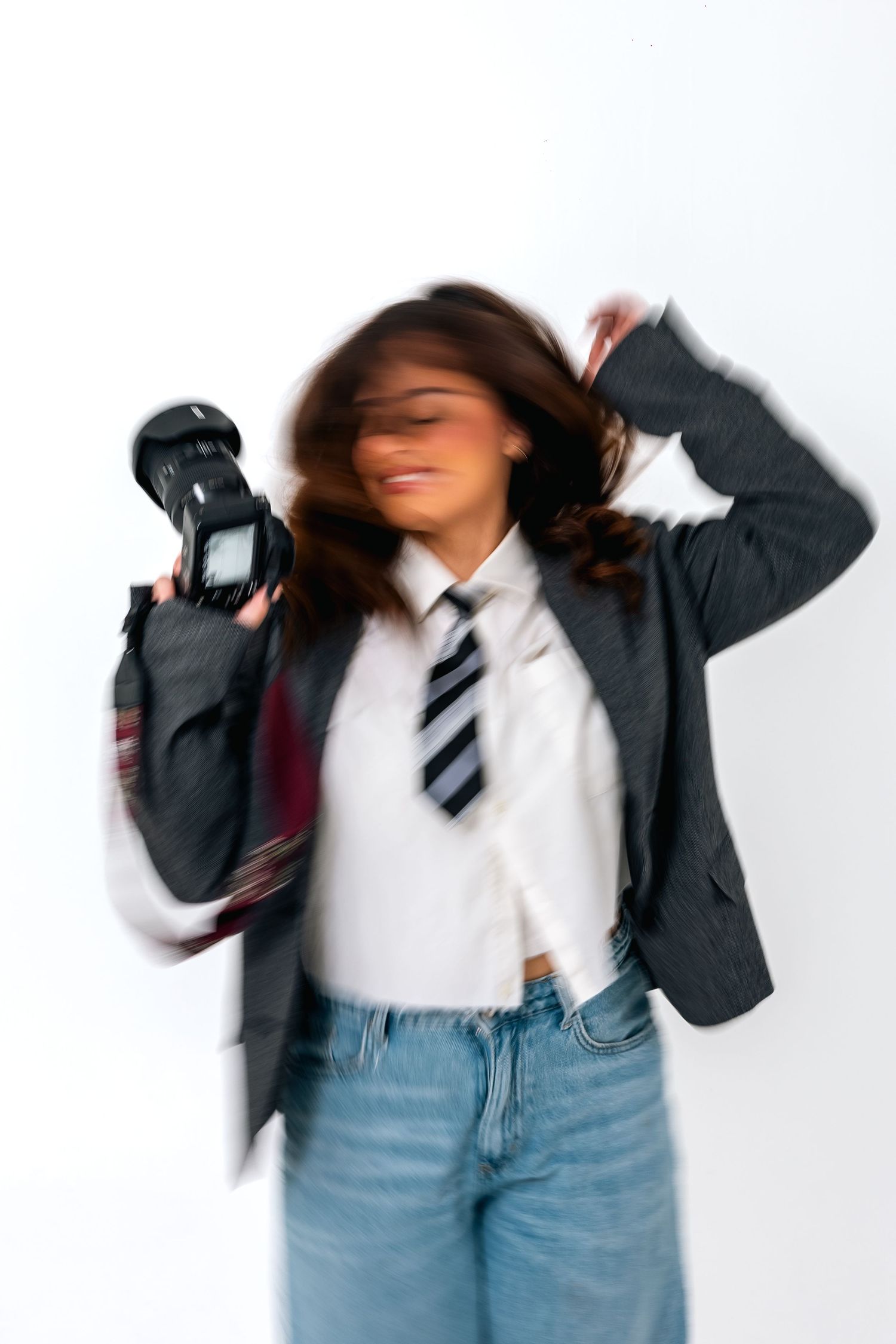 Blurred motion shot of a young woman in blazer and tie holding a DSLR camera against a white background.
