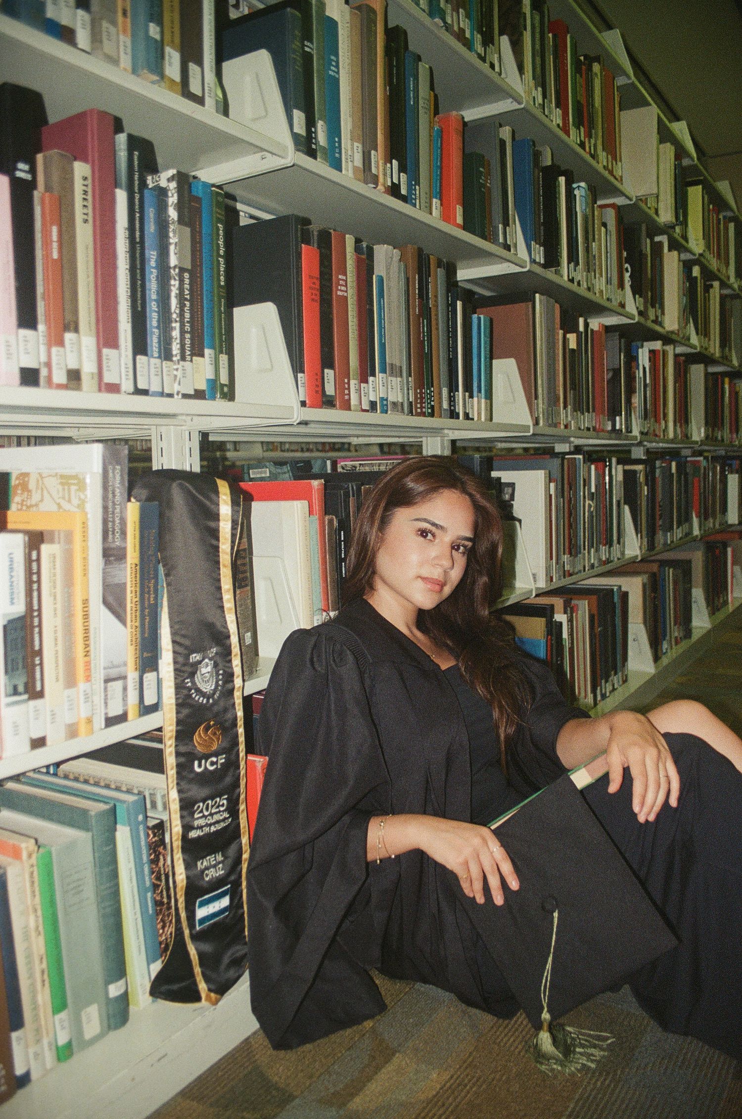 Female graduate in black gown sits beside library bookshelves with UCF cap and stole.