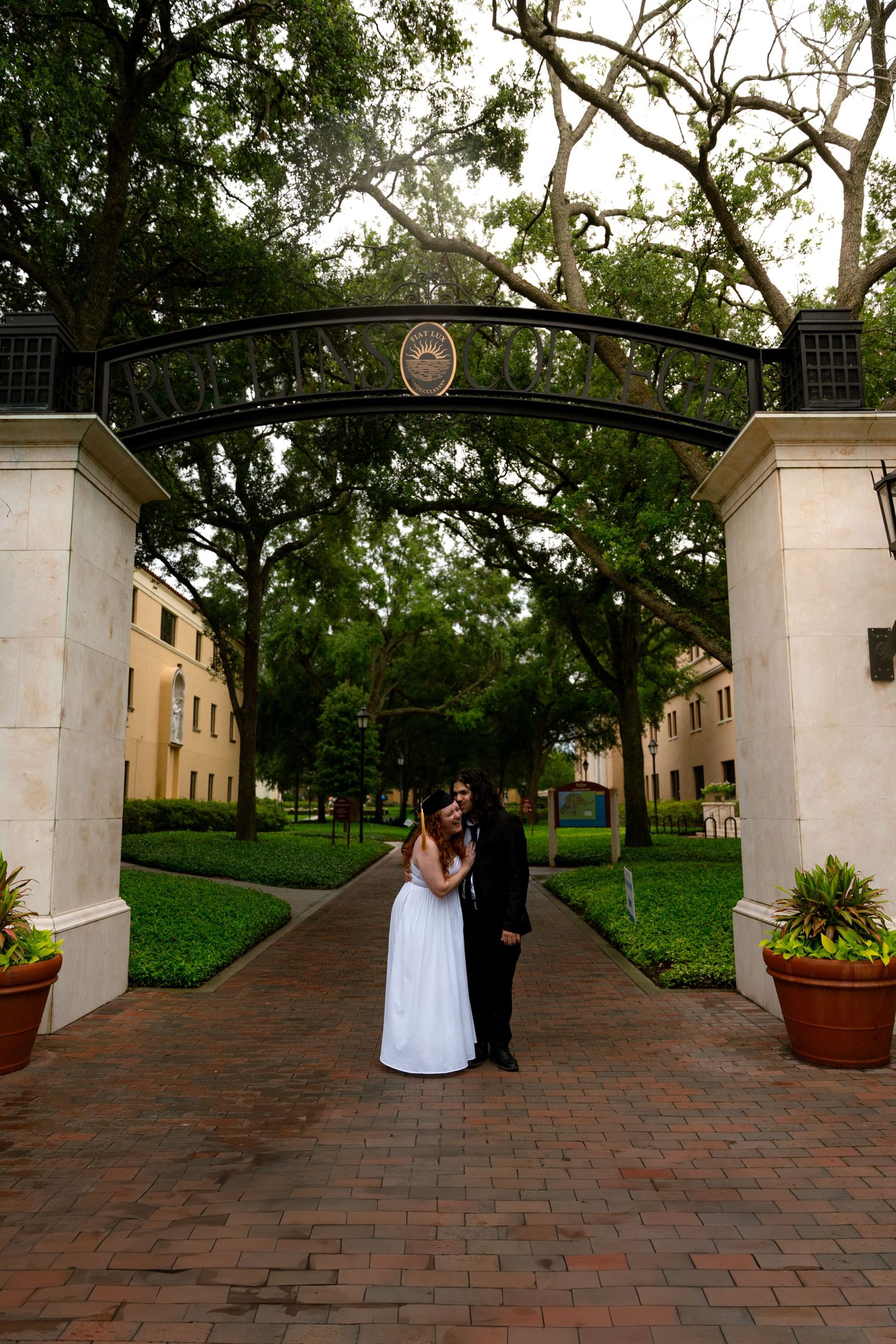 Bride and groom pose under ornate iron archway on brick path surrounded by lush green trees at elegant venue.