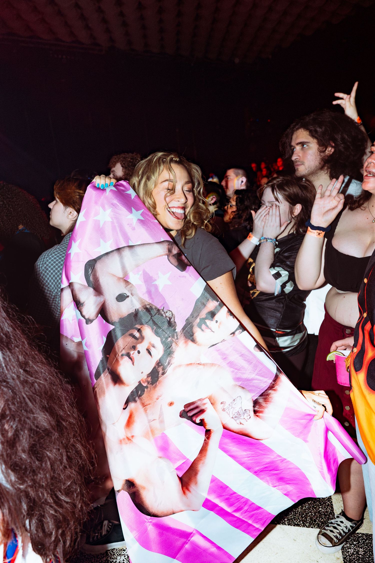 Excited fan holds a large artist flag while smiling at a crowded indoor concert.