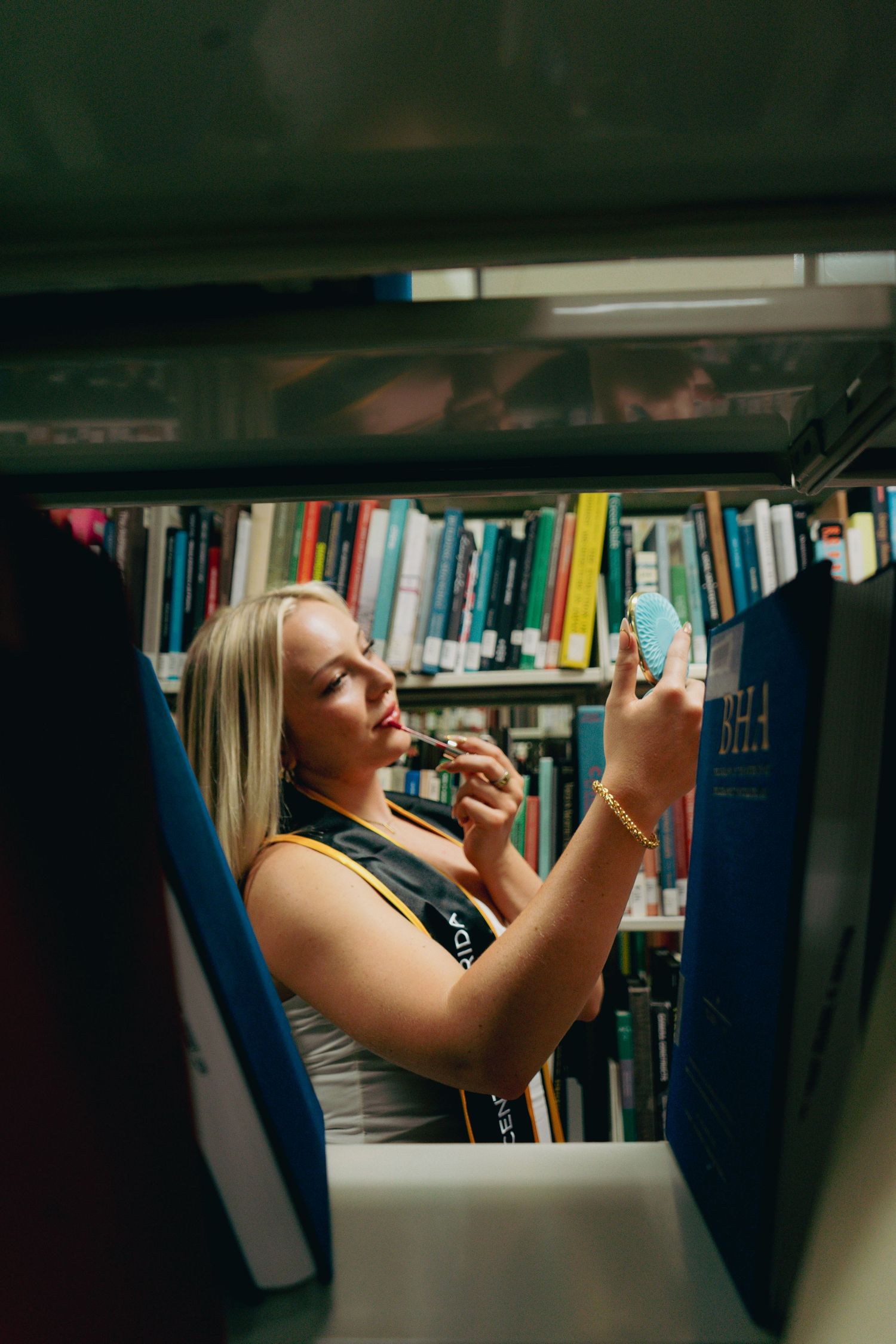 Blonde graduate in cap and gown applies lipstick using compact mirror between library bookshelves.