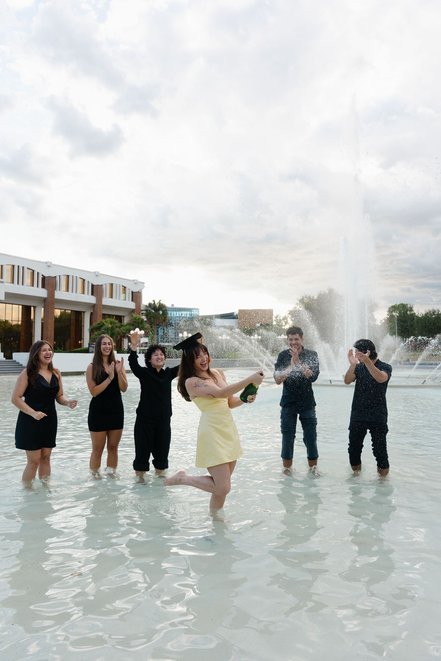 Group of friends celebrating in a fountain, popping champagne in formal attire on a cloudy day.