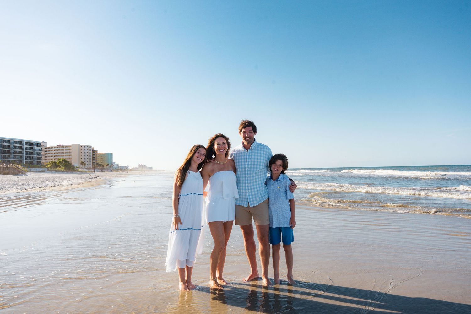 Family of four smiling together on a sunny beach with clear blue sky and ocean waves behind them.