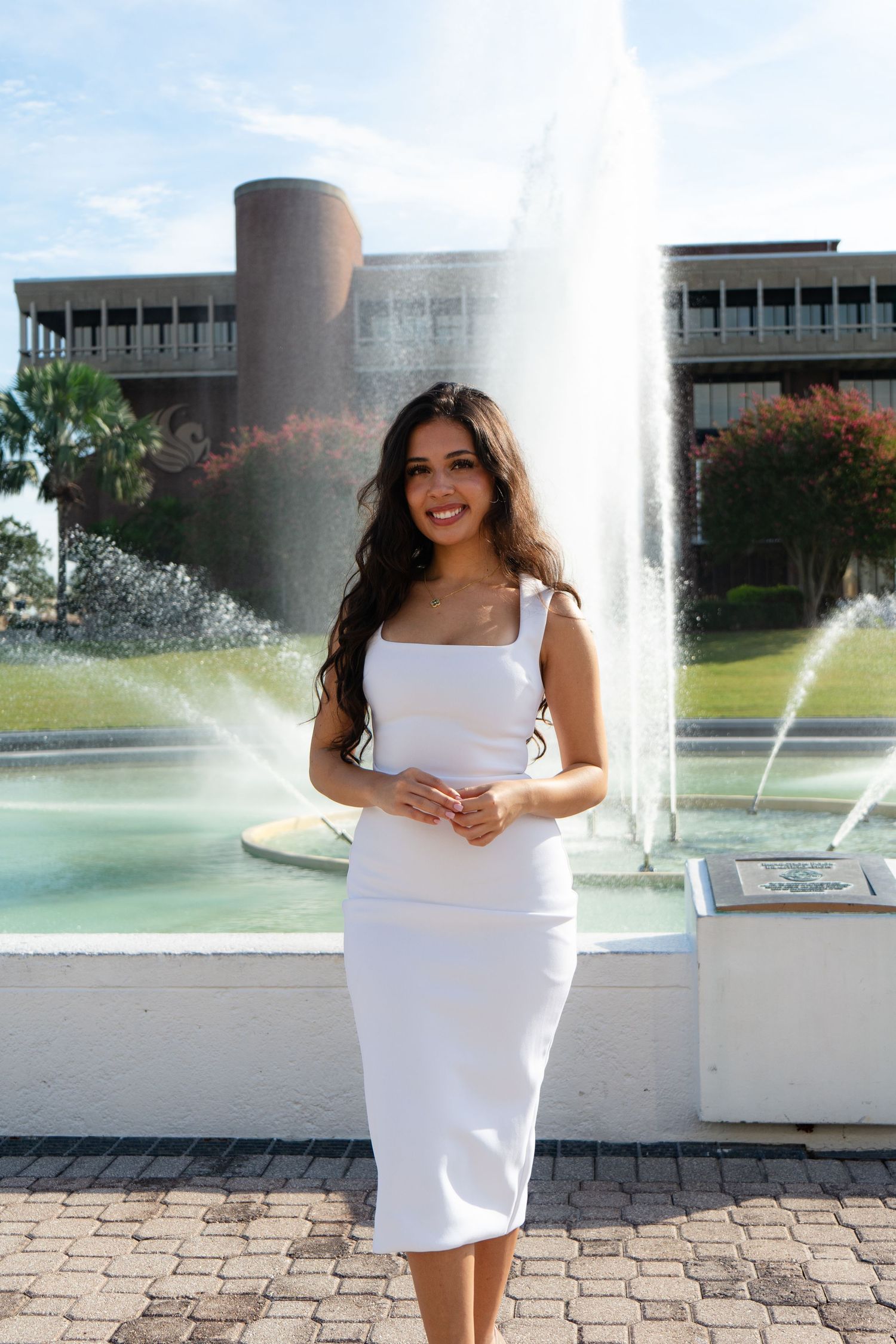 Young woman in white midi dress smiling in front of a fountain on a sunny day.