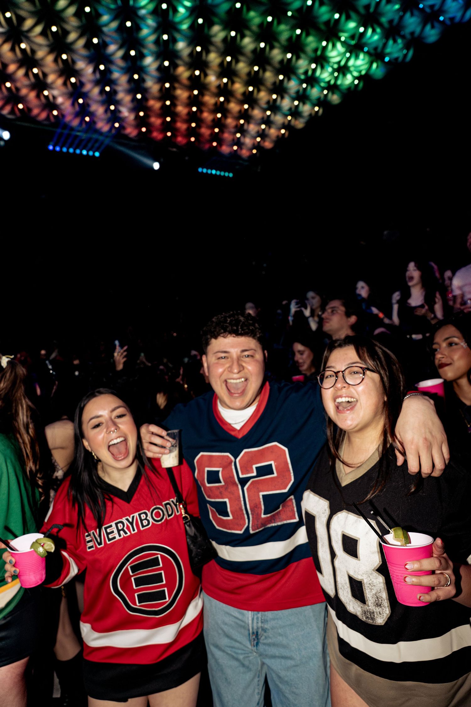 Three friends in hockey jerseys smiling and holding drinks under colorful lights at a concert.